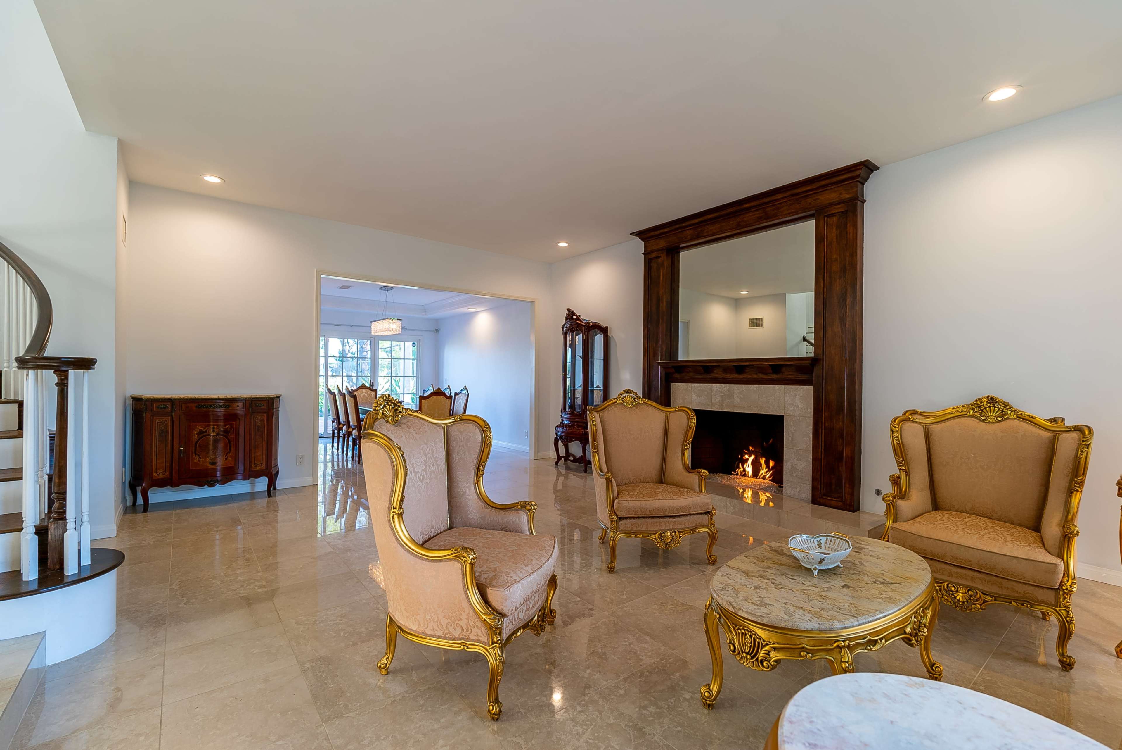 A living room with two ornate chairs, a marble-topped coffee table, and a fireplace, connected to a dining area visible through an archway.