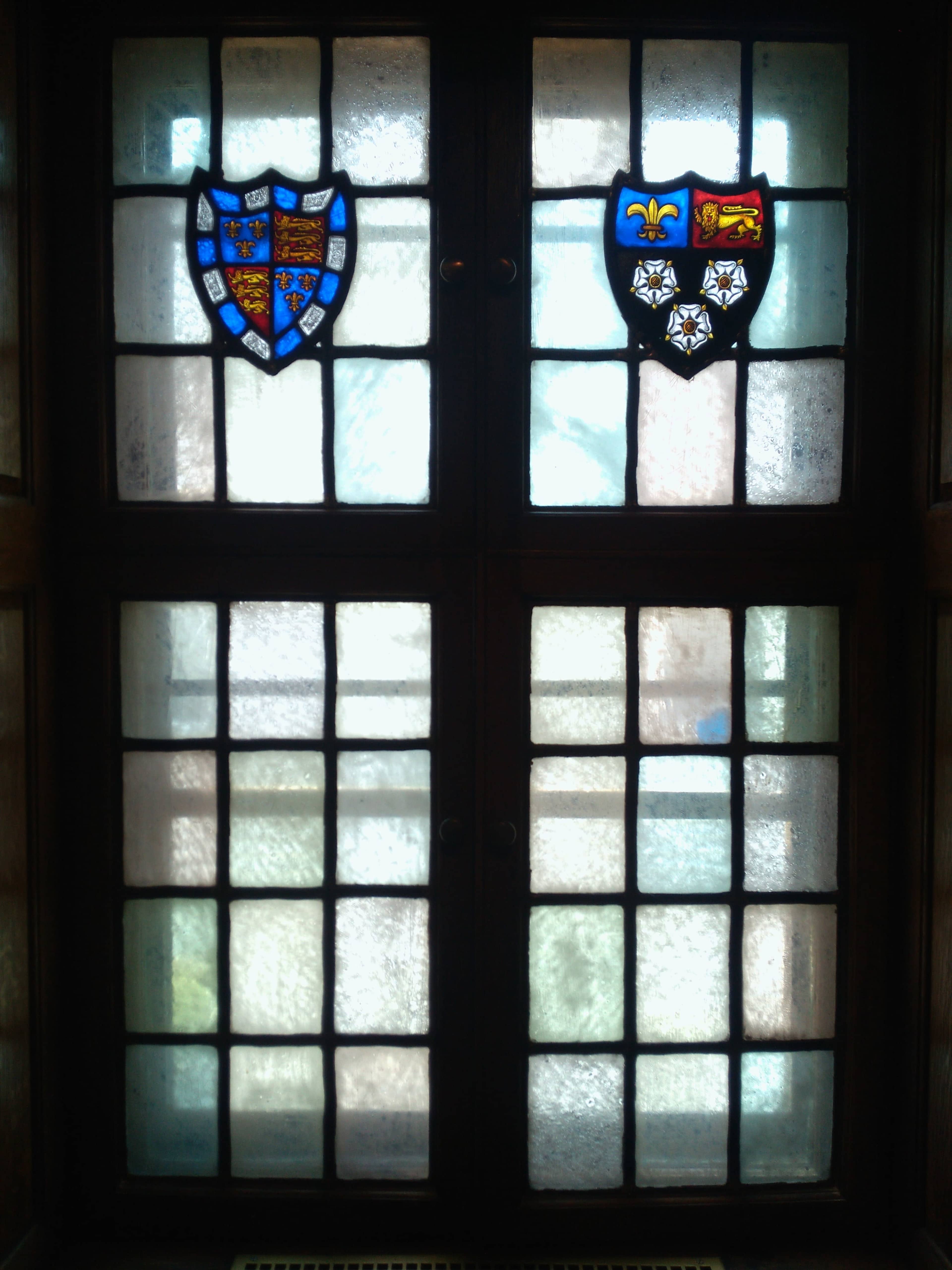 The image shows a window with two stained glass coats of arms, framed by a grid of wooden window panes.