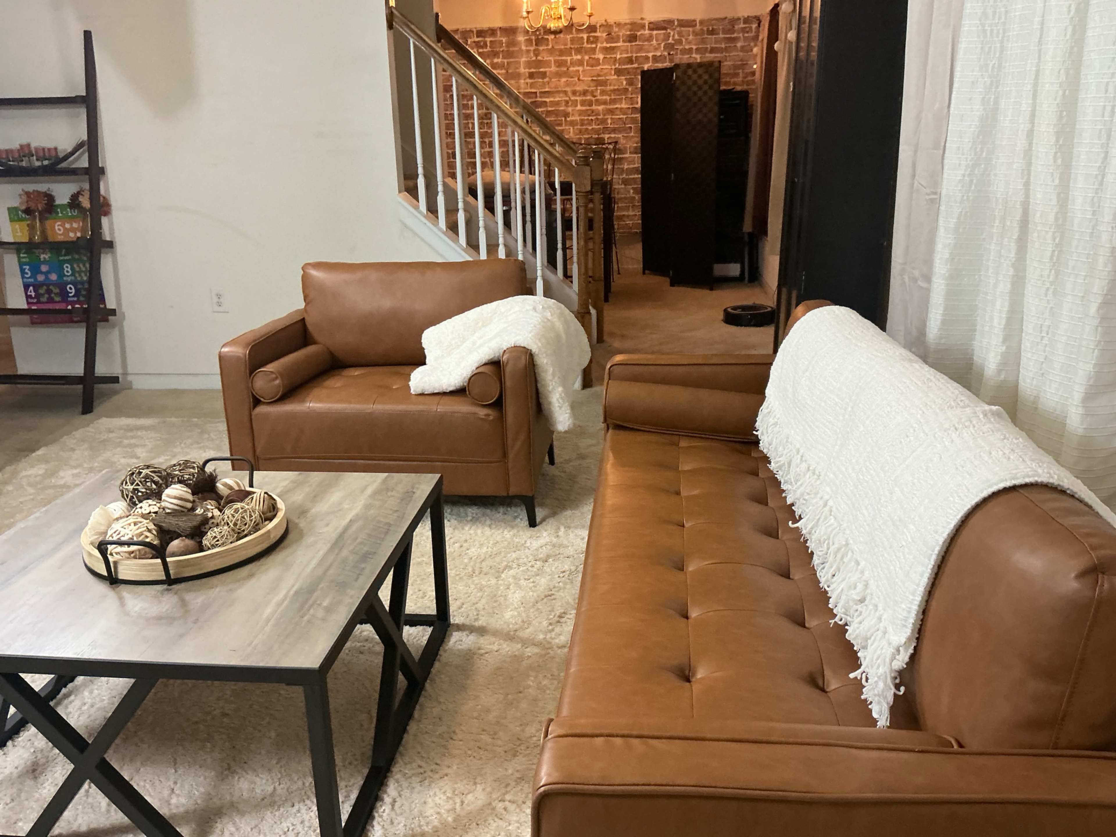 The scene features a cozy living area with two brown leather armchairs, a tan sofa, a wooden coffee table with a decorative bowl, and a light-colored rug.