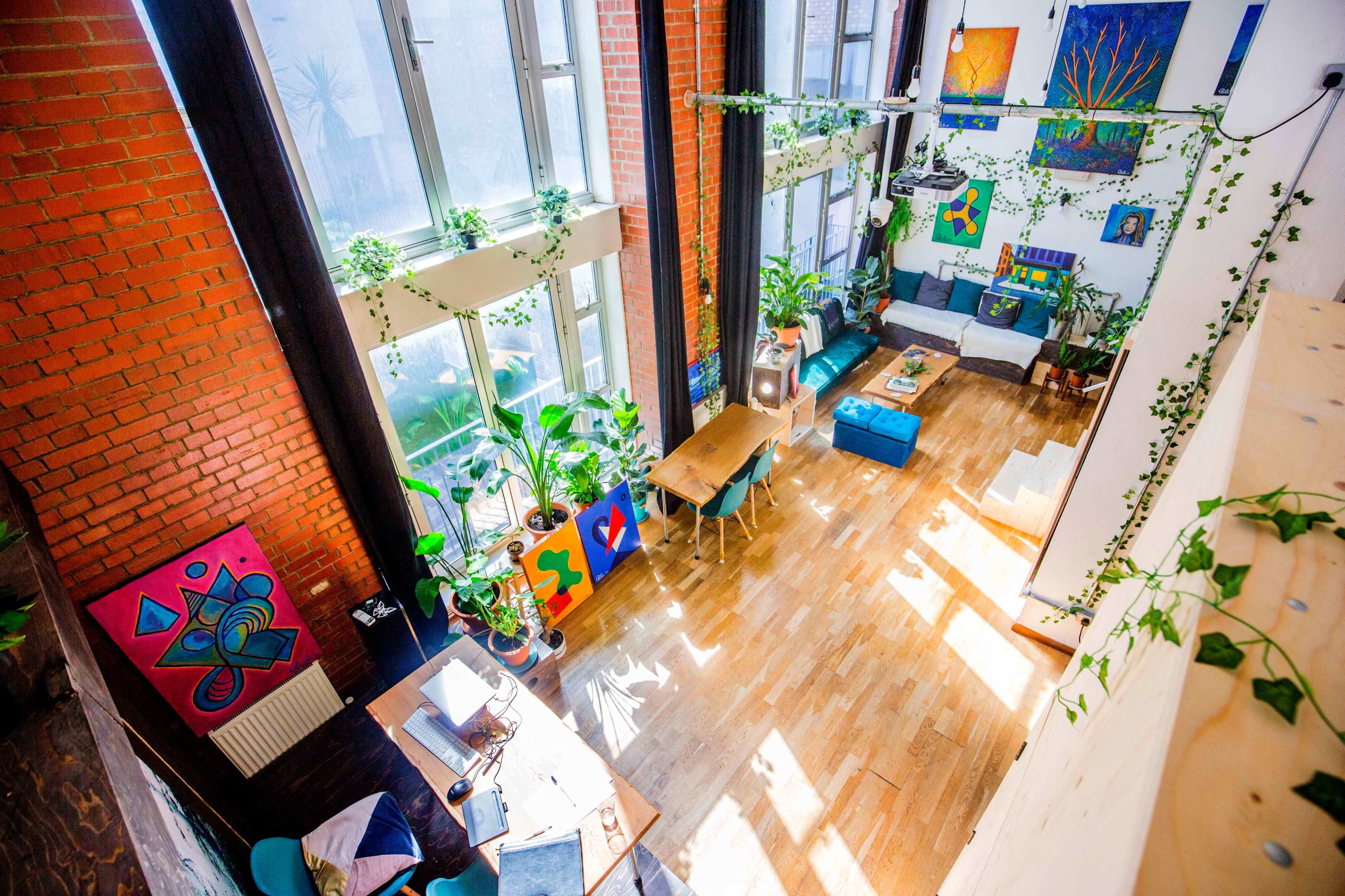 A bright and spacious room with large windows, wooden floors, plants, and colorful artwork on the walls.