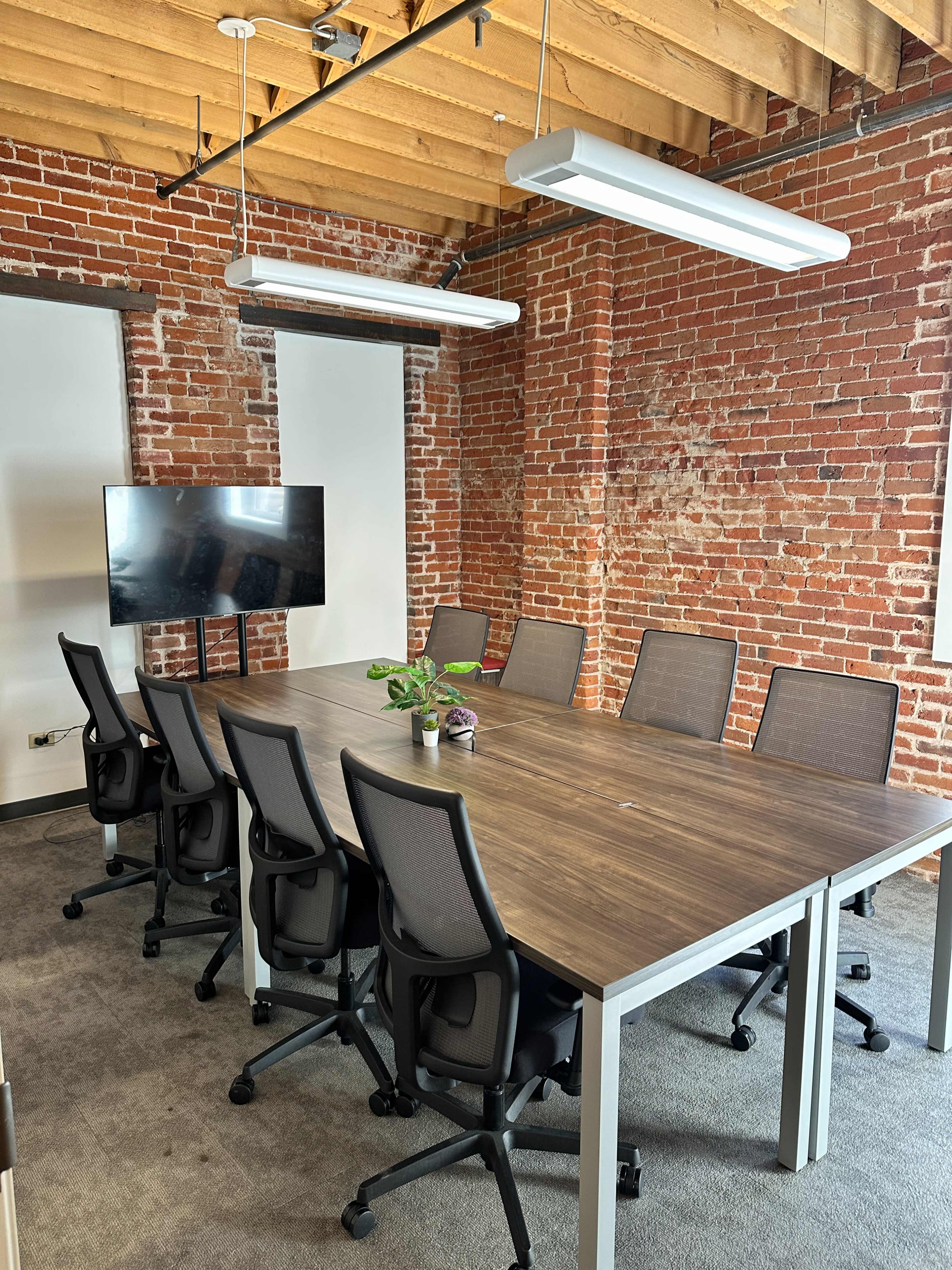 The image shows a modern conference room with a large wooden table surrounded by ergonomic chairs, exposed brick walls, and a television mounted on the wall.