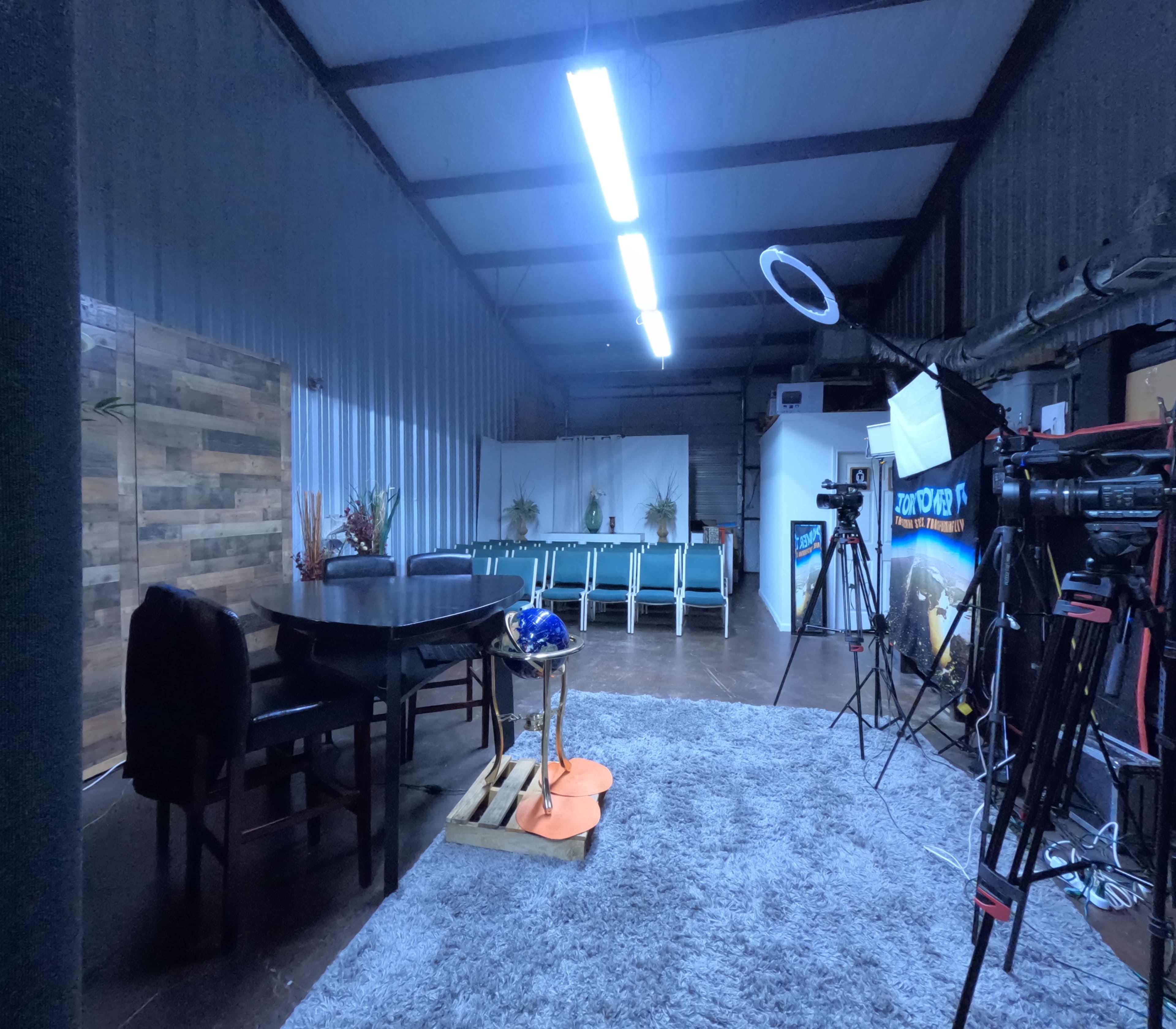 The image shows a spacious room with a dining table and chairs, a set of faux plants, and filming equipment positioned around the area.