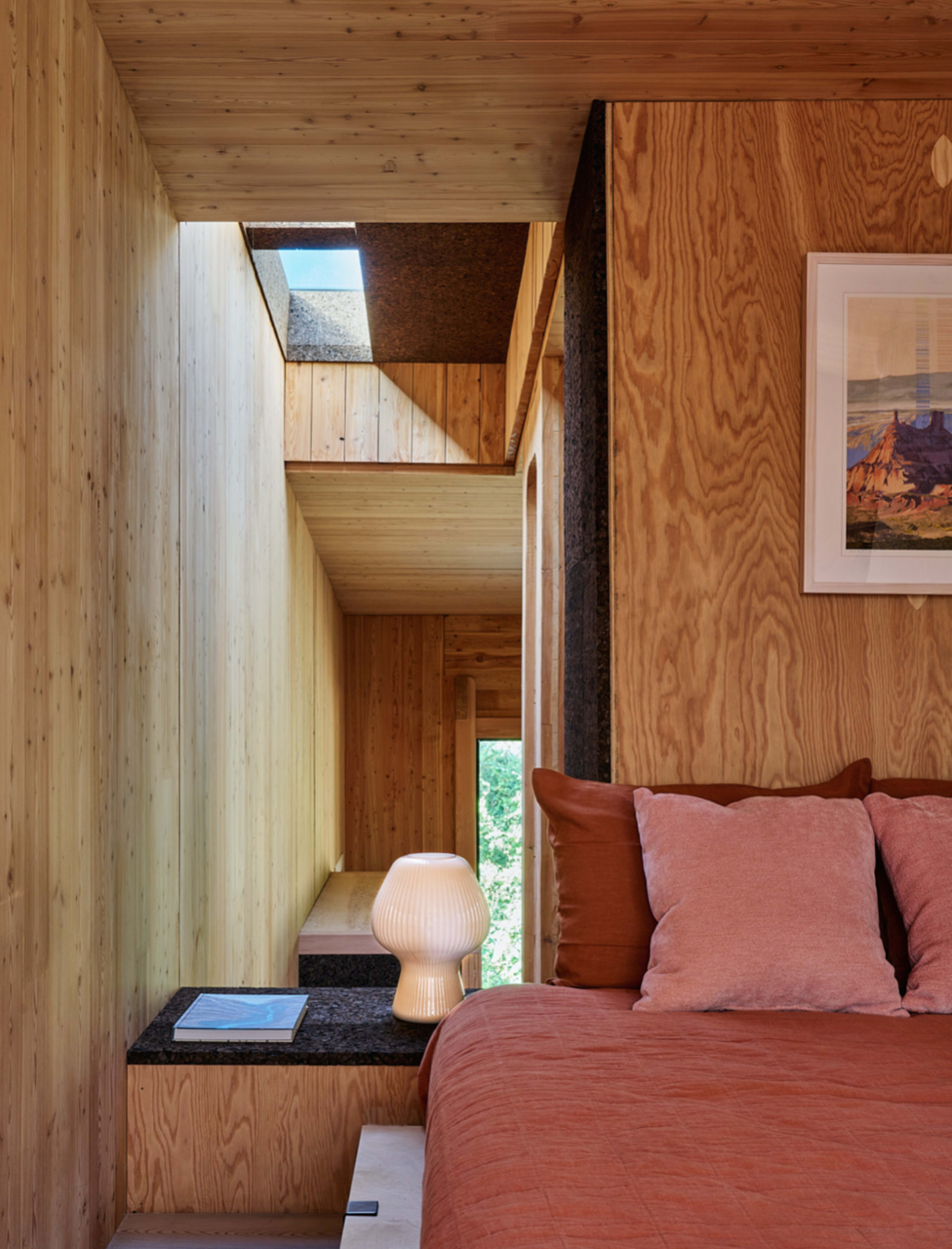 A modern bedroom with wooden walls, a cozy bed with orange bedding, a lamp on a bedside table, and a narrow hallway leading to a skylight.