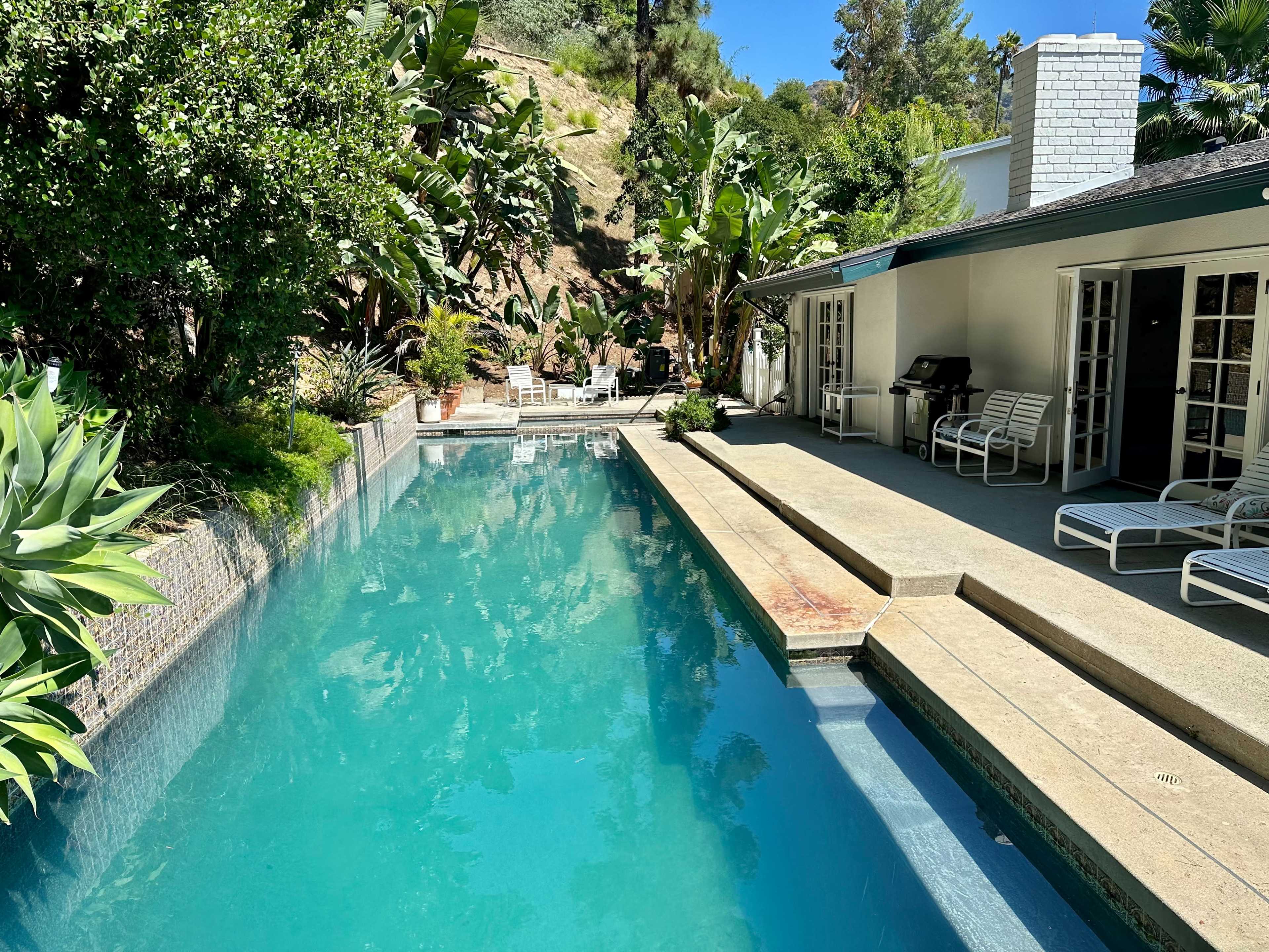 The image shows a clear swimming pool surrounded by lush greenery and a patio area with lounge chairs.