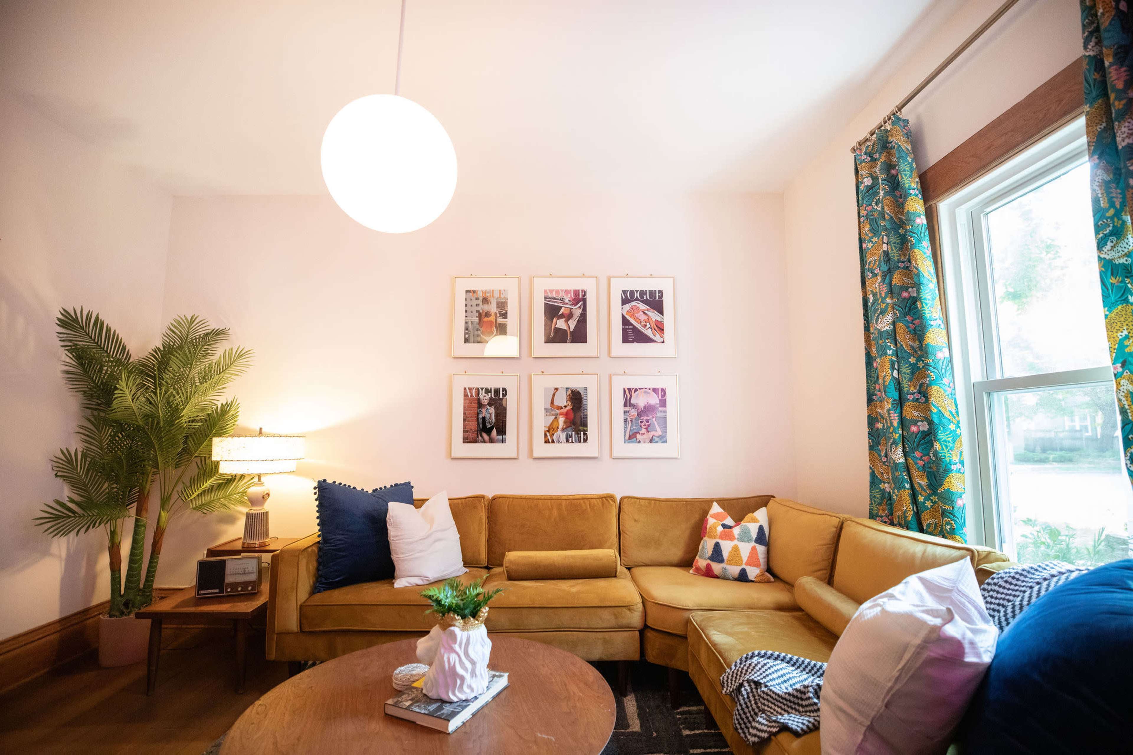 A cozy living room features a yellow sectional sofa, a round coffee table, decorative pillows, and framed artwork on the wall, with a floor lamp and a potted plant in the corner.