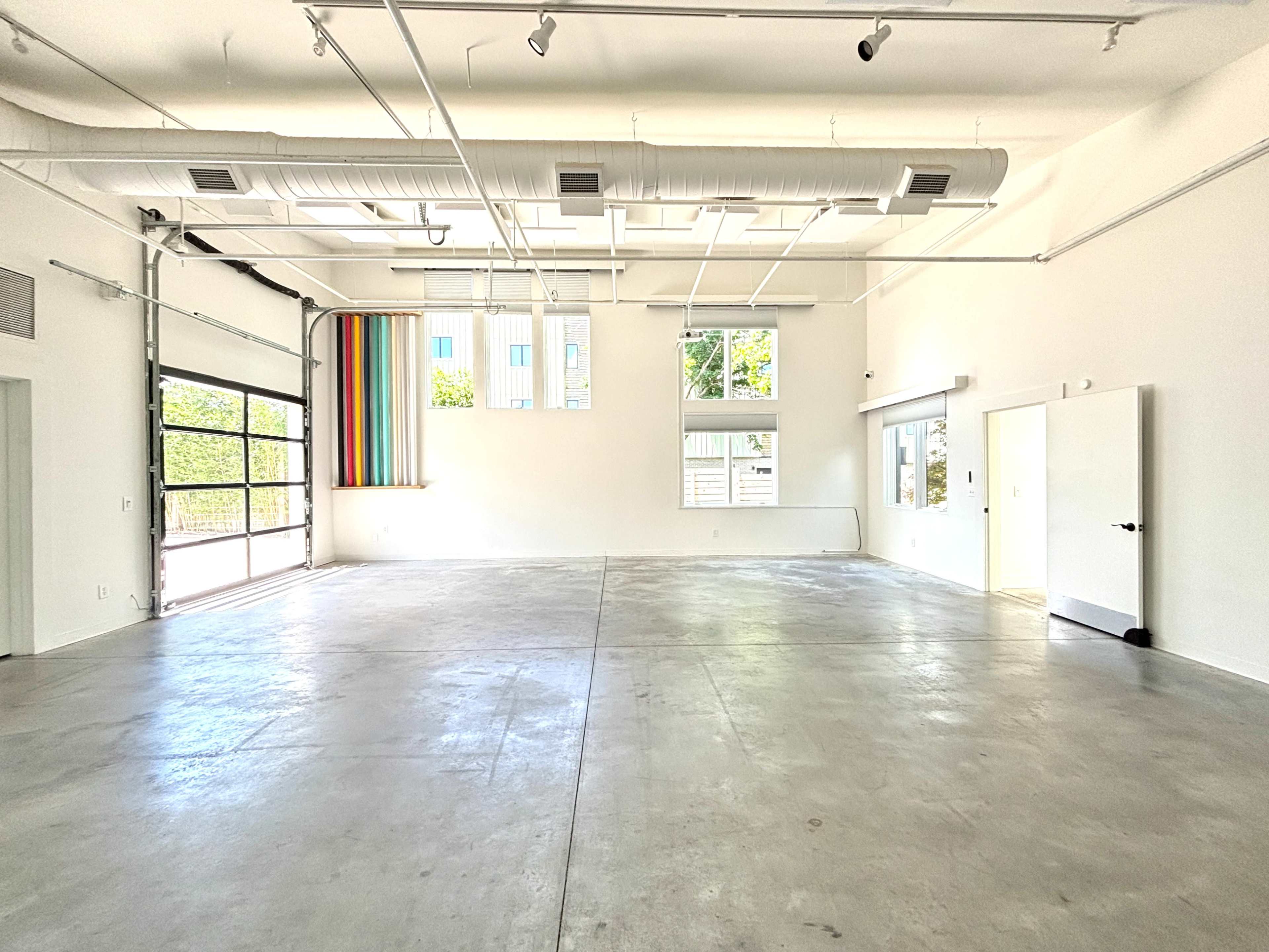 The image shows a spacious, empty room with high ceilings, a large garage door, and colorful window treatments on one side.