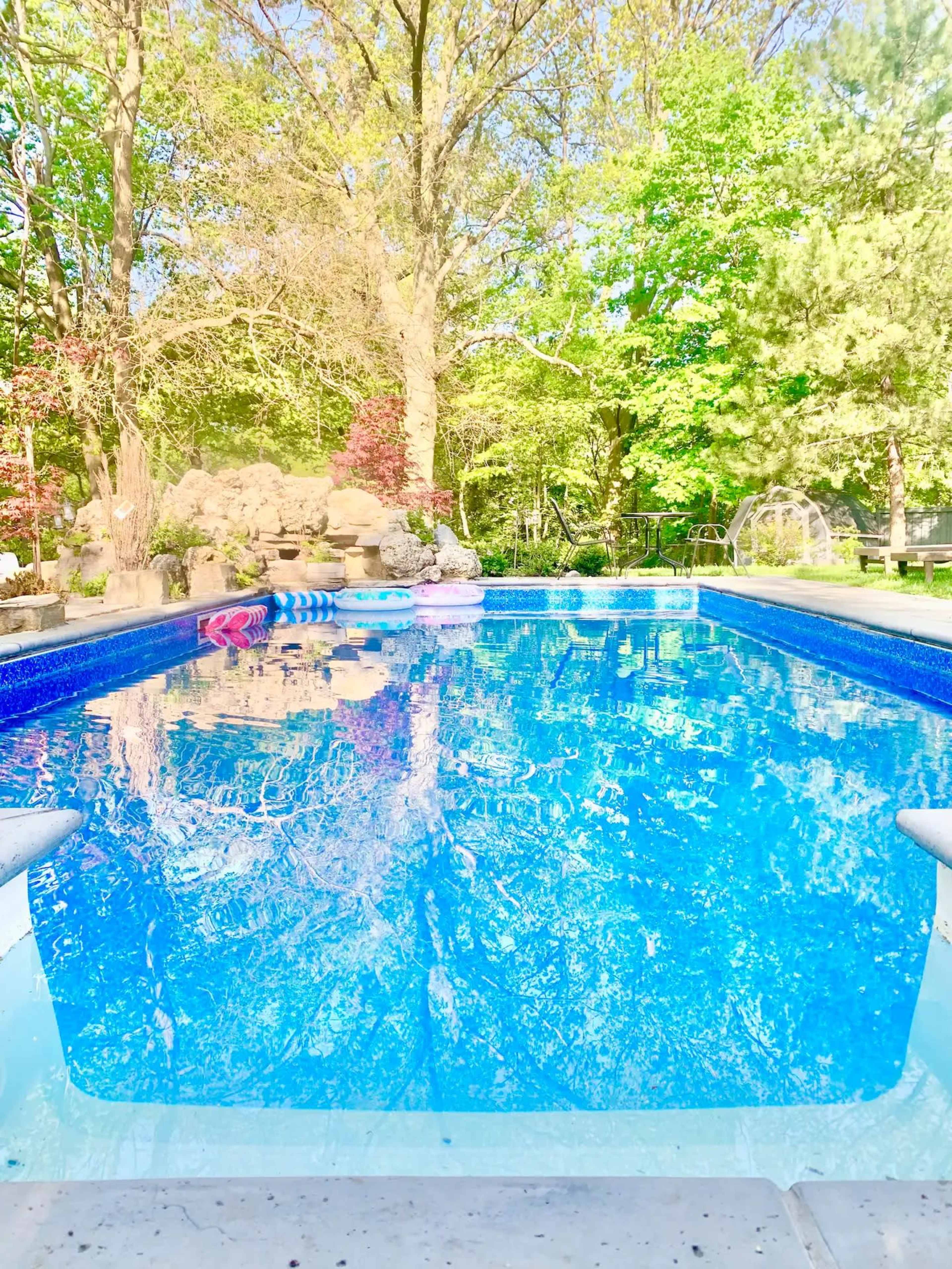 A clear, blue swimming pool is surrounded by trees and a stone feature in a backyard setting.