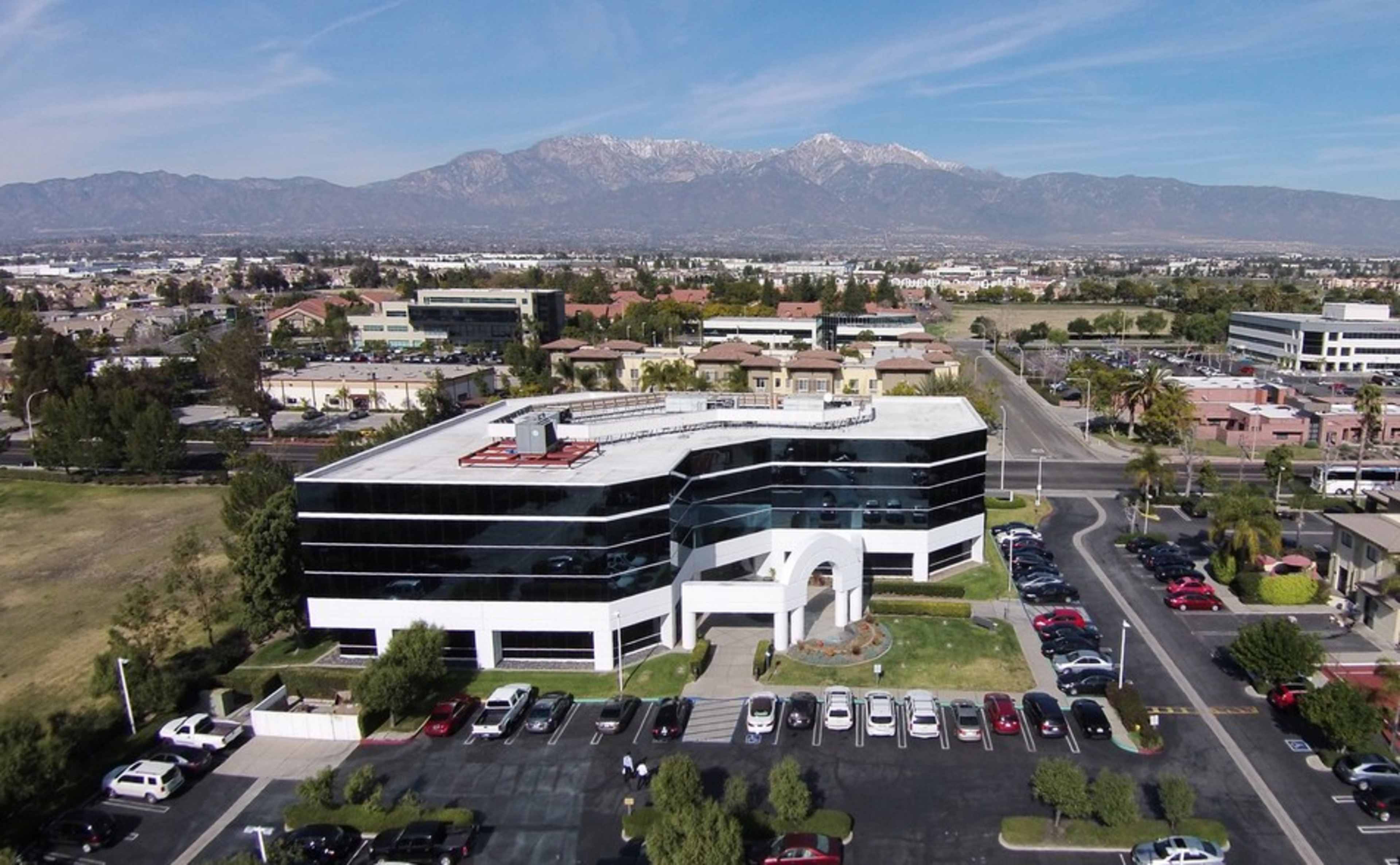 The image shows a modern office building with large glass windows surrounded by a parking lot and mountains in the background.
