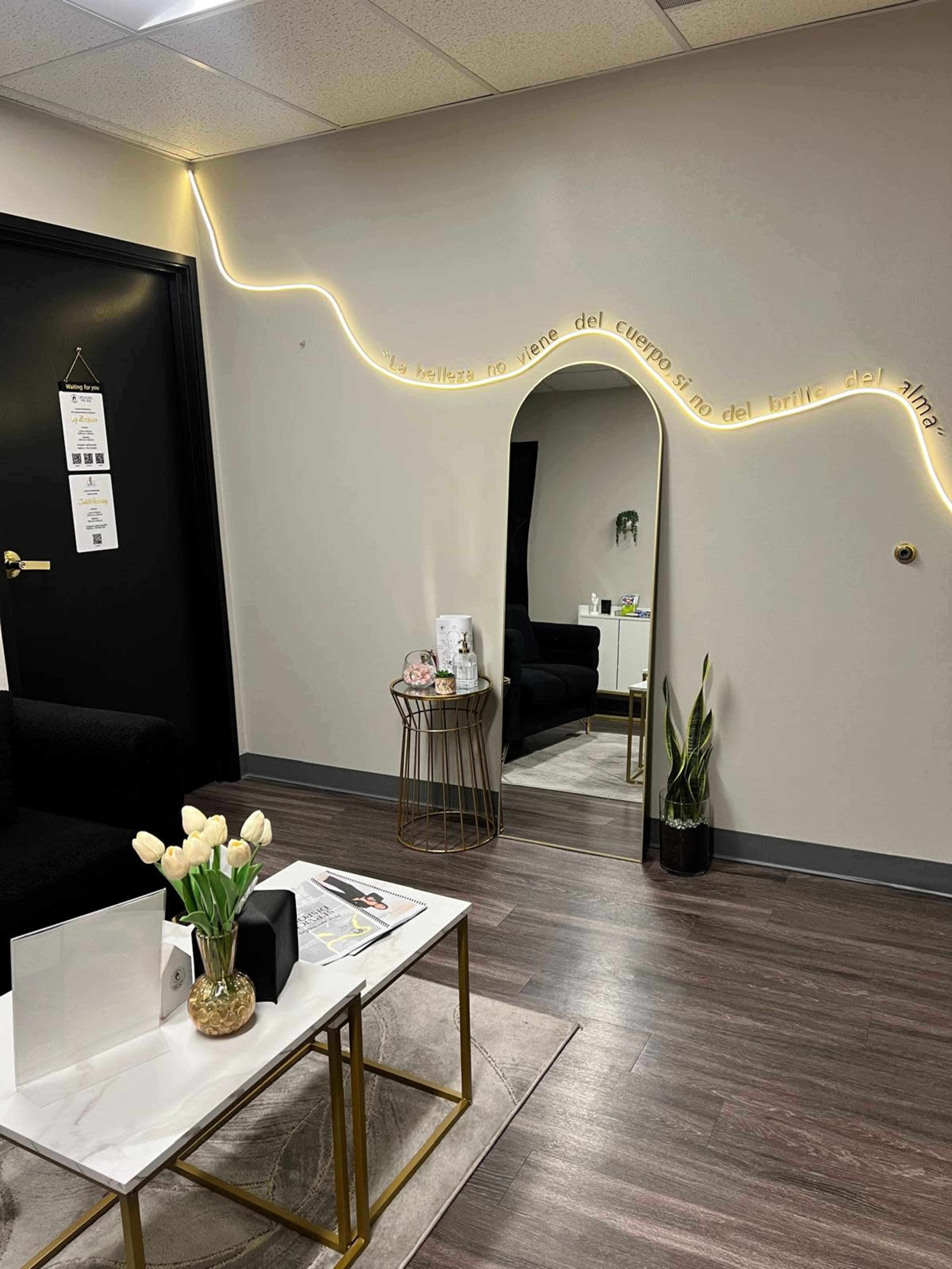 The image shows a modern waiting area with a black sofa, a mirror, a small table with flowers, and ambient lighting along the wall.
