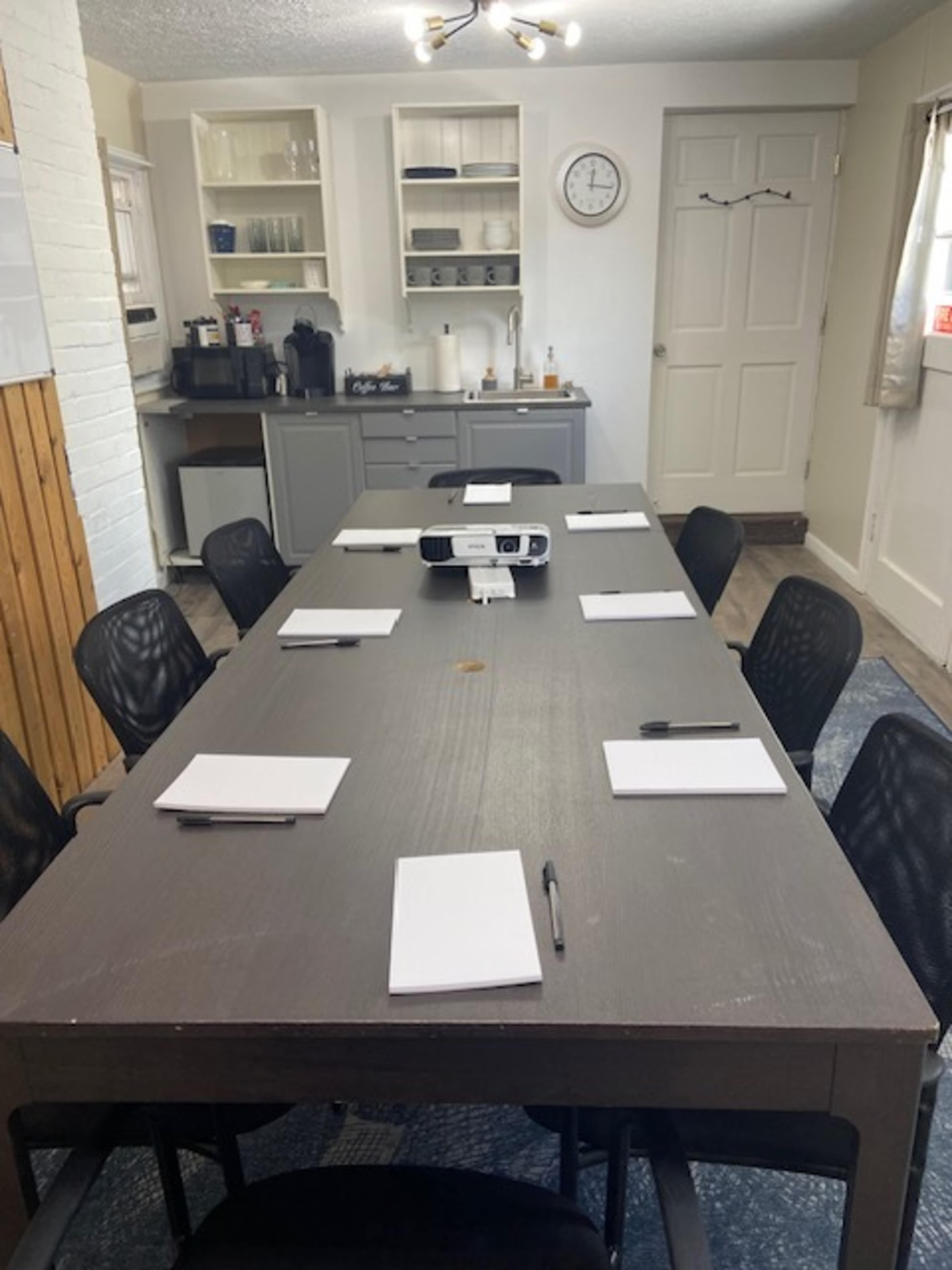 A long conference table with pads and pens is set up in a bright room featuring a coffee station and a clock on the wall.