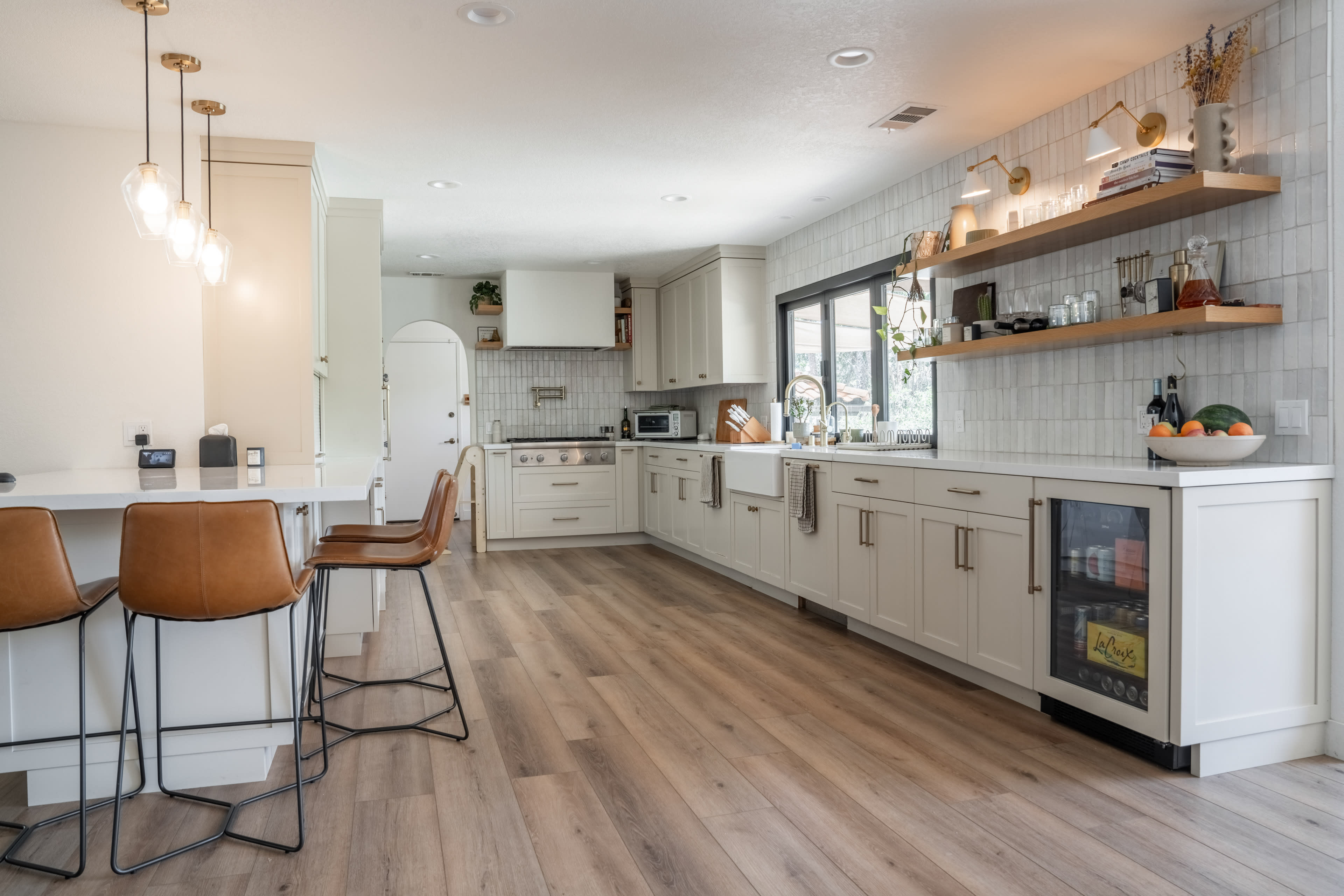 The image shows a modern kitchen with light cabinetry, wooden flooring, a central island with bar stools, and a window above the sink.