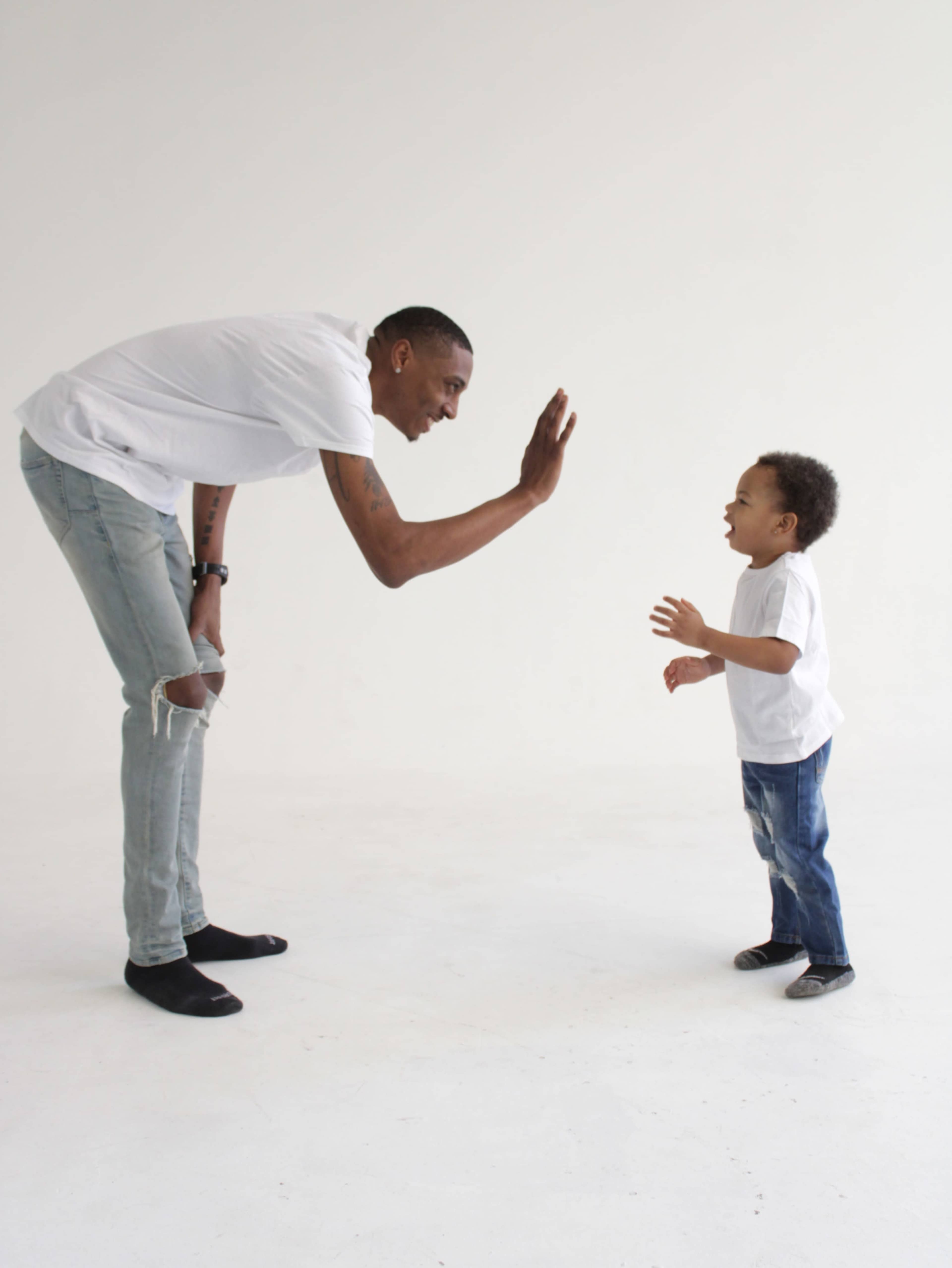 A man and a young child are facing each other, with the man raising his hand as if to greet the child, who is smiling and reaching out.