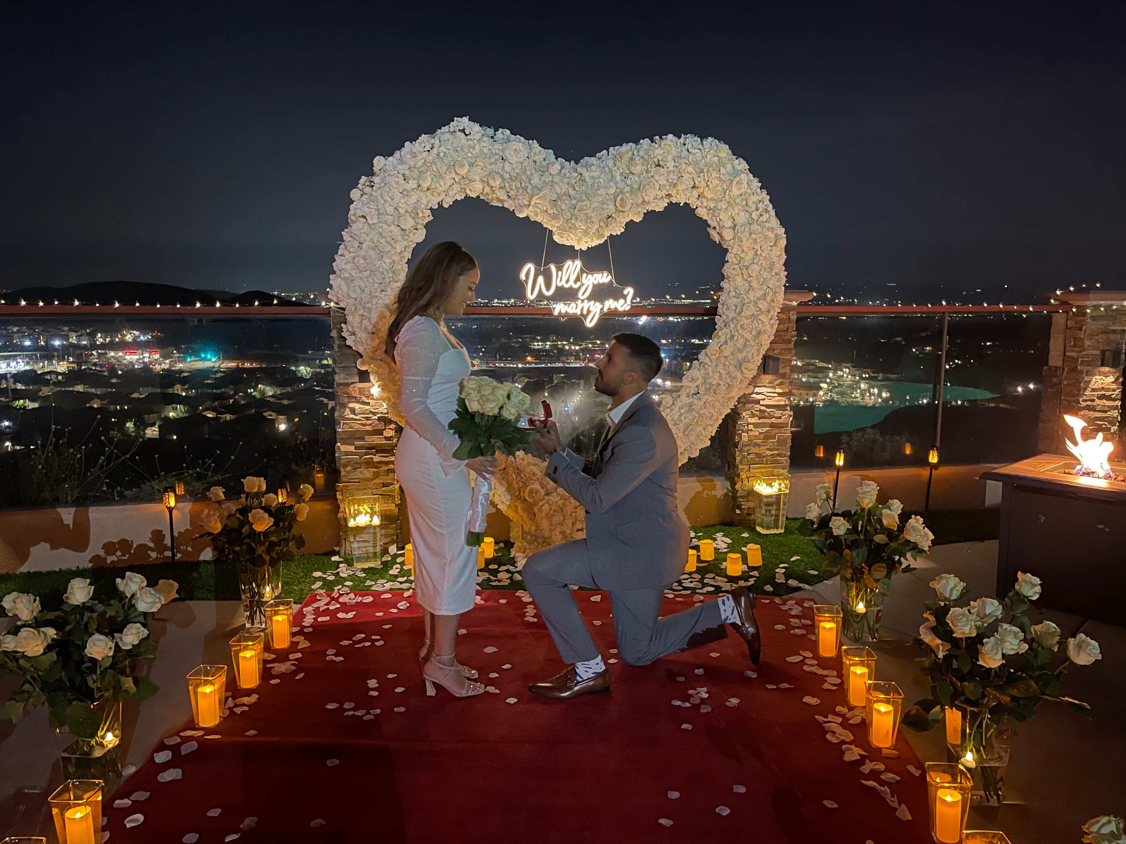 A man is proposing to a woman in front of a heart-shaped floral display, surrounded by candles and a city skyline at night.