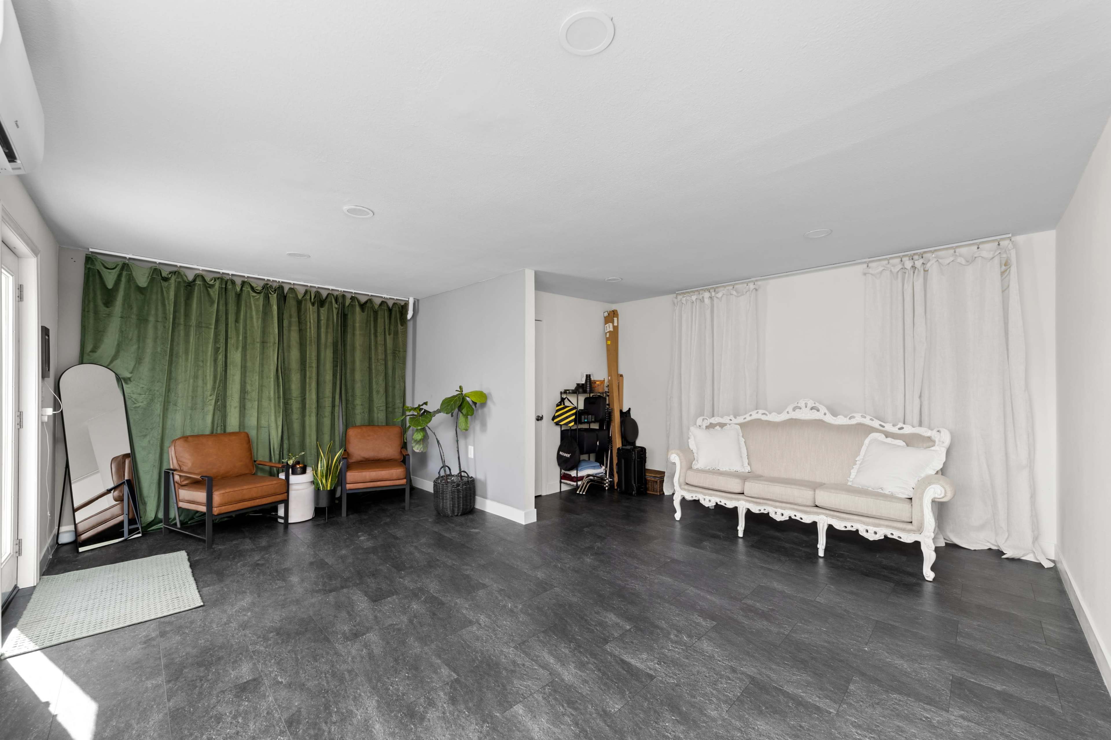 The image shows a spacious room with two brown chairs, a white sofa, a mirror, and green curtains along one wall.