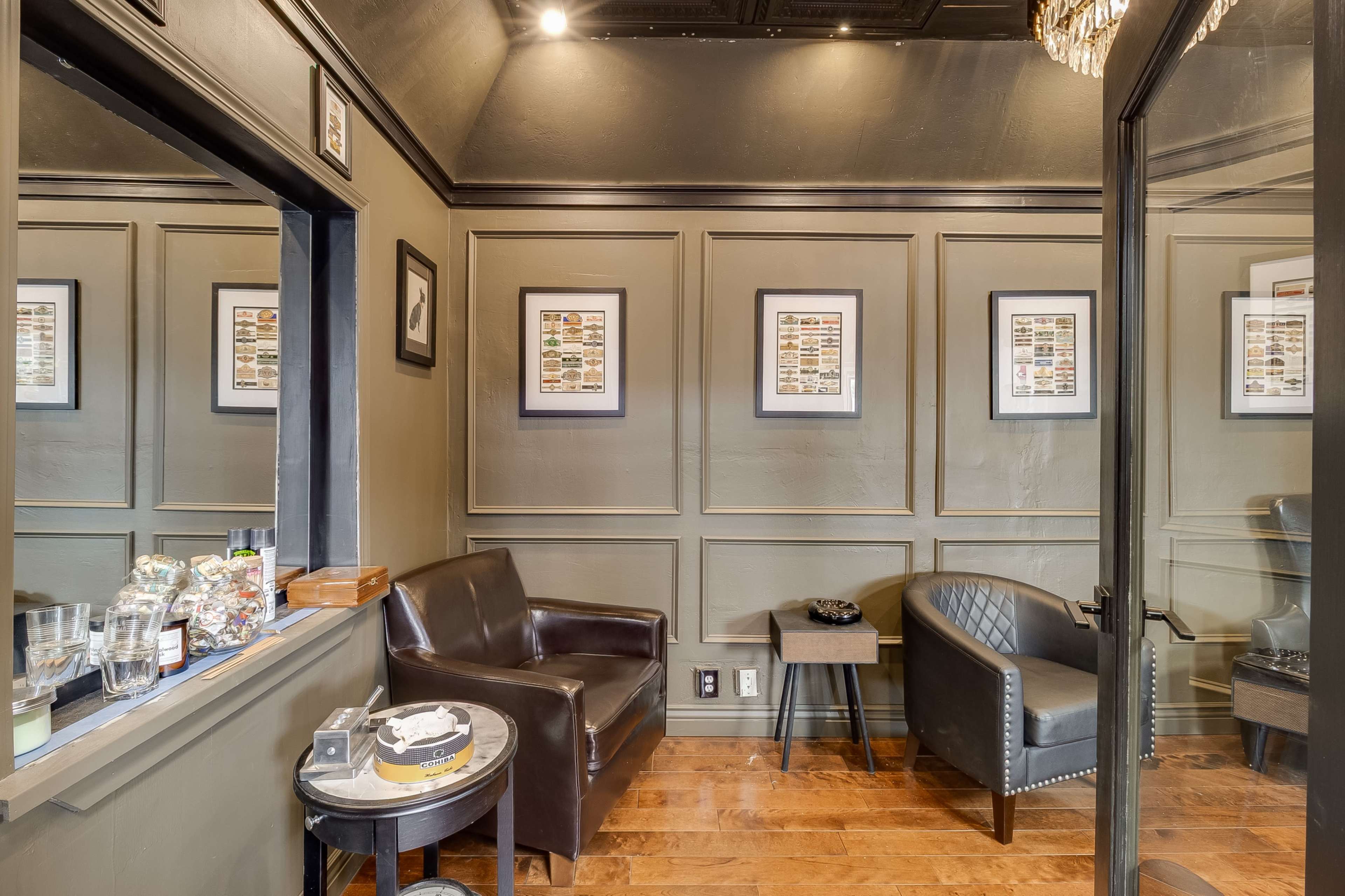 The image shows a small, elegantly designed room with dark green walls, featuring two chairs, framed artwork, and a glass door.