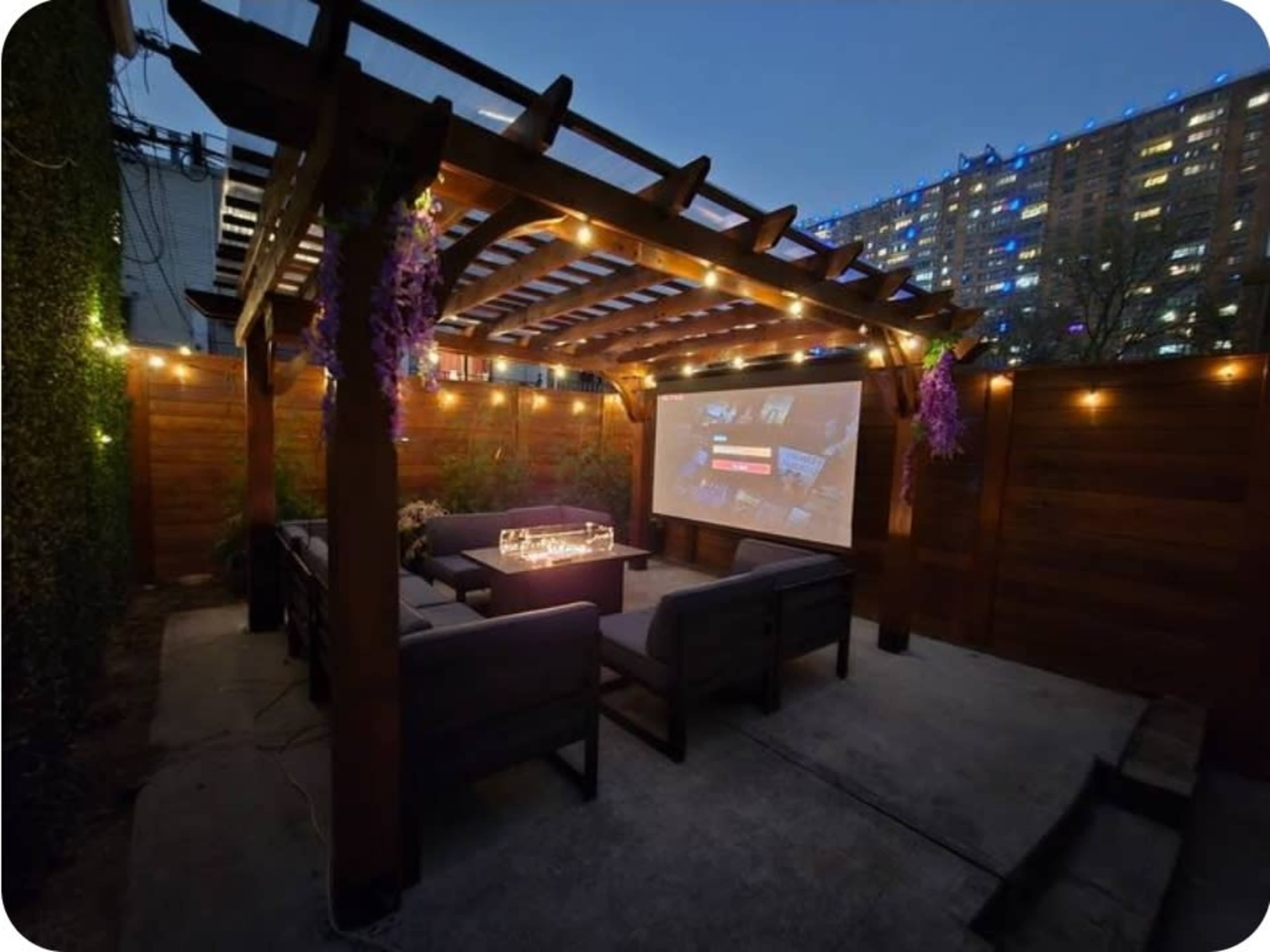 The image shows an outdoor patio with a wooden pergola, string lights, and a large screen displaying a movie or presentation, surrounded by urban buildings in the background.