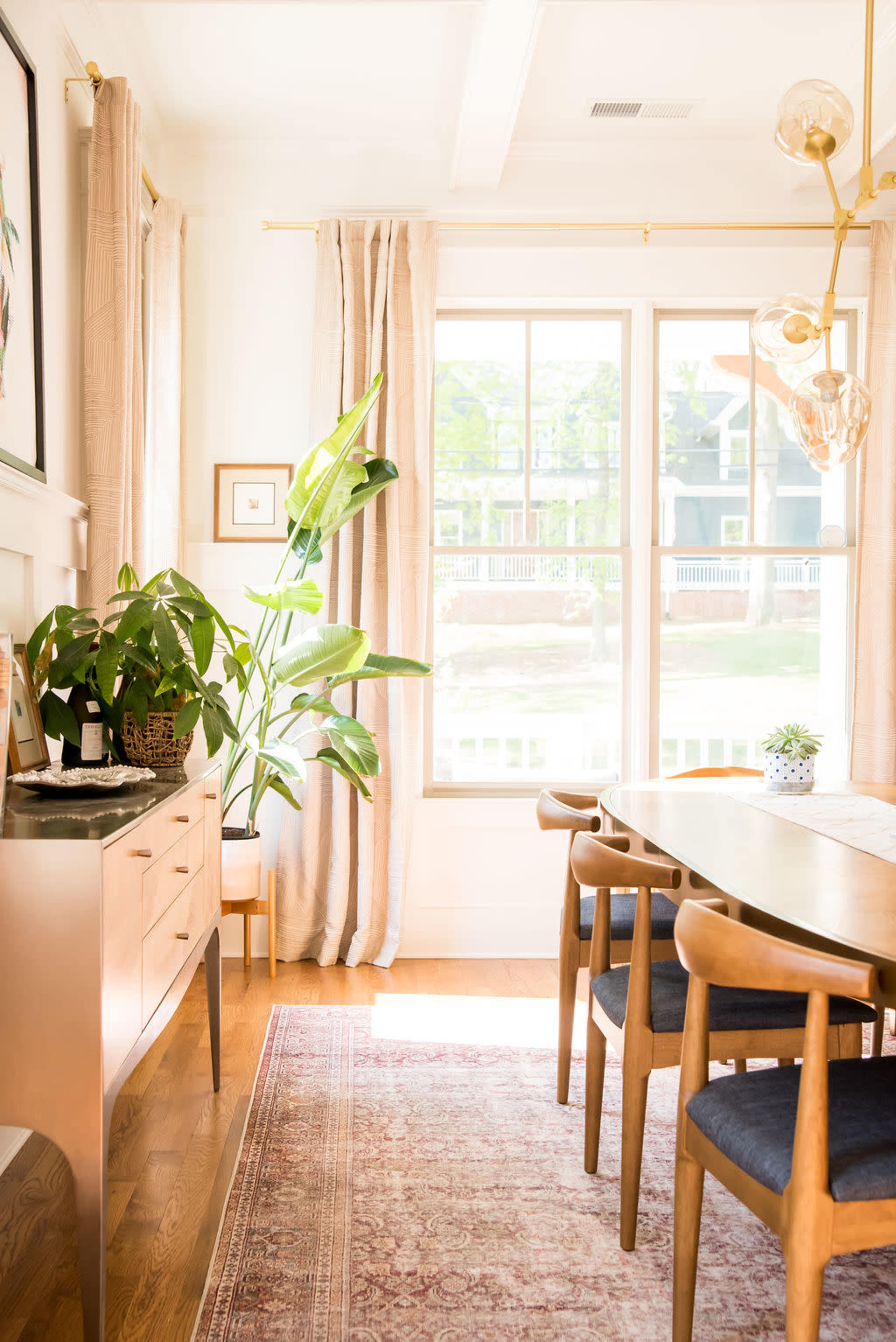A dining room features a wooden table with modern chairs, a sideboard, a large window with curtains, and a houseplant in the corner.