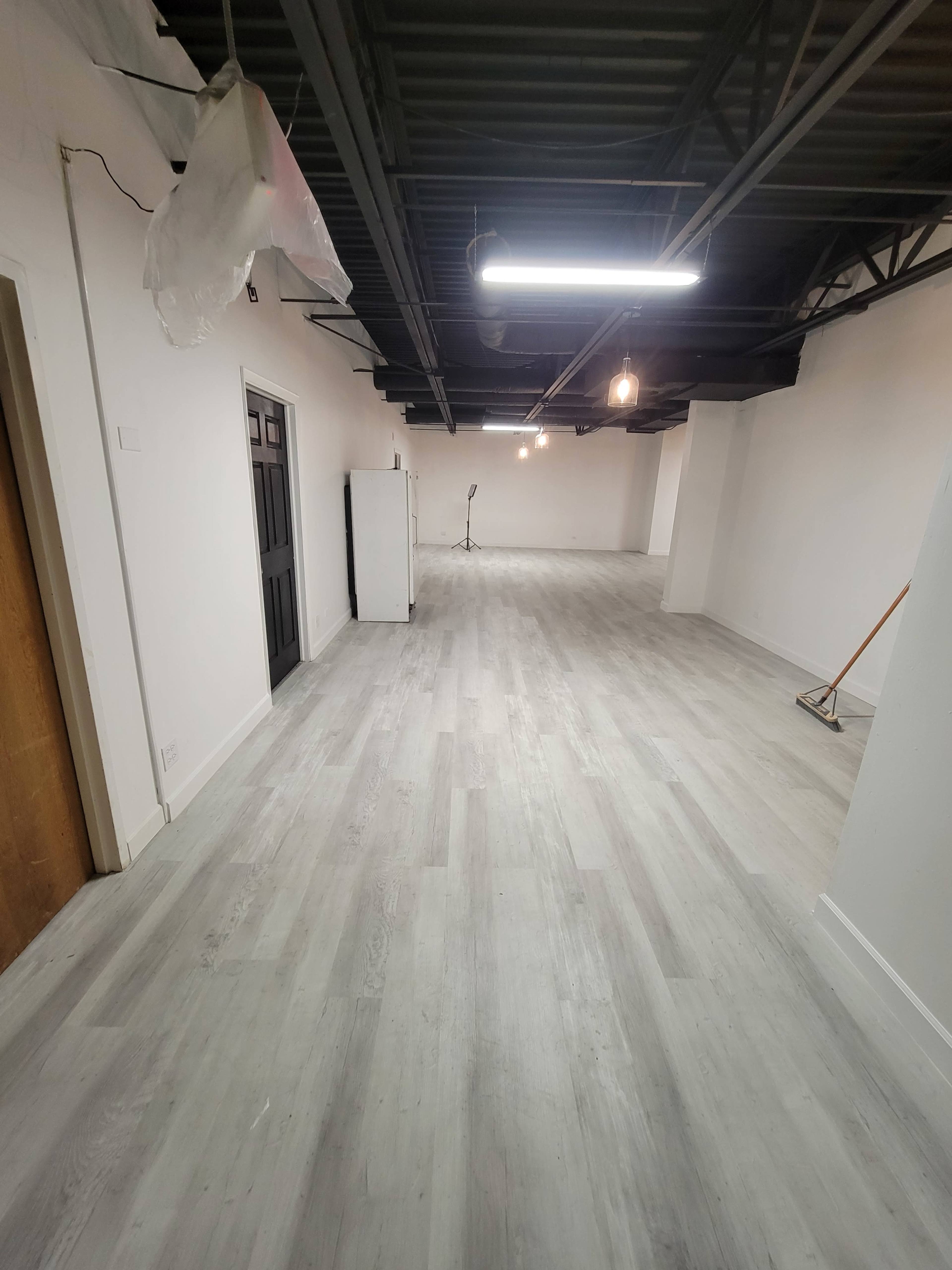 A spacious, unfinished room with light gray flooring, white walls, and exposed ceiling beams, illuminated by overhead lights.
