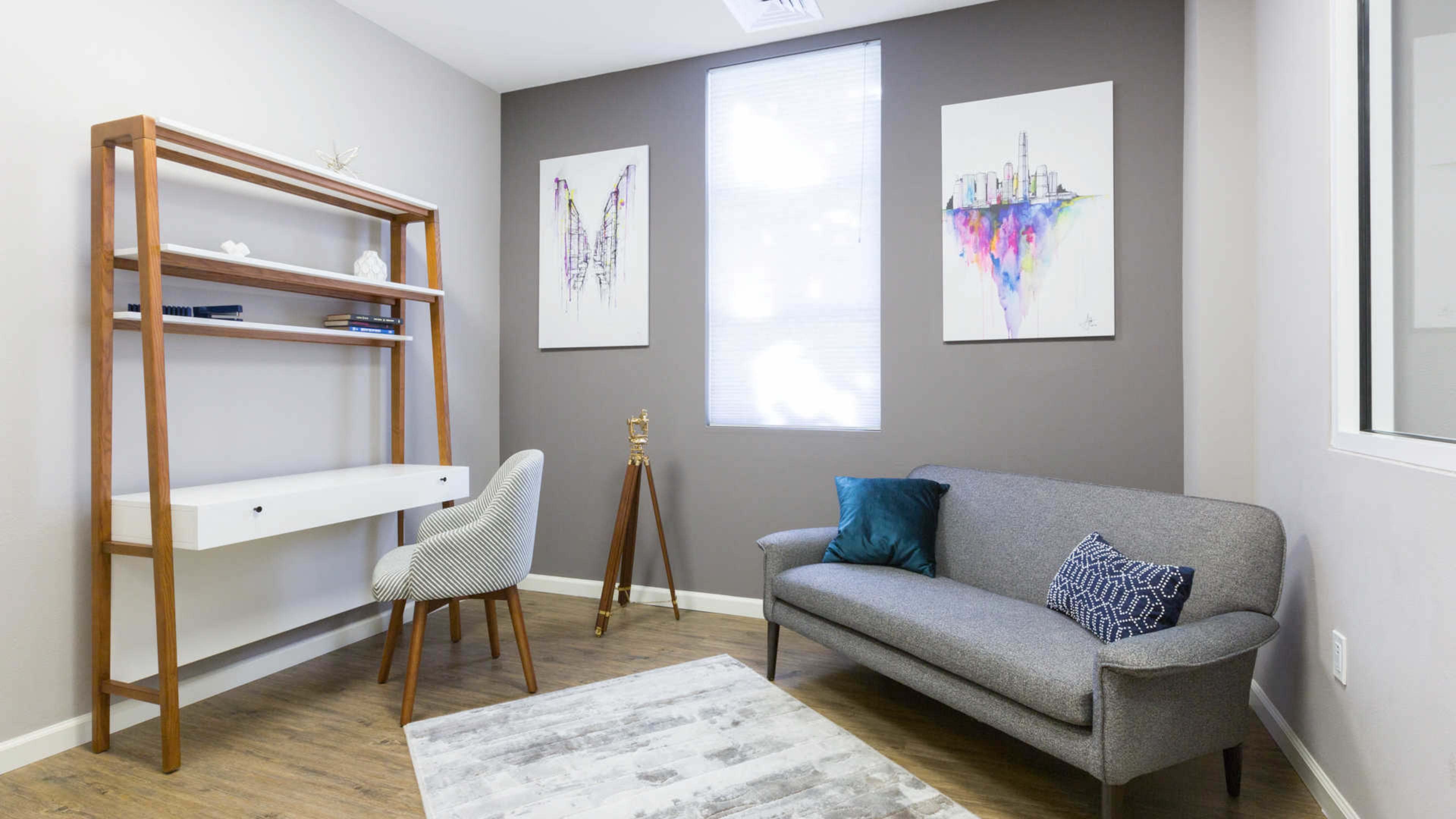 The image shows a modern, minimalistic office space featuring a gray sofa, a wooden bookshelf, and two abstract art pieces on the wall.