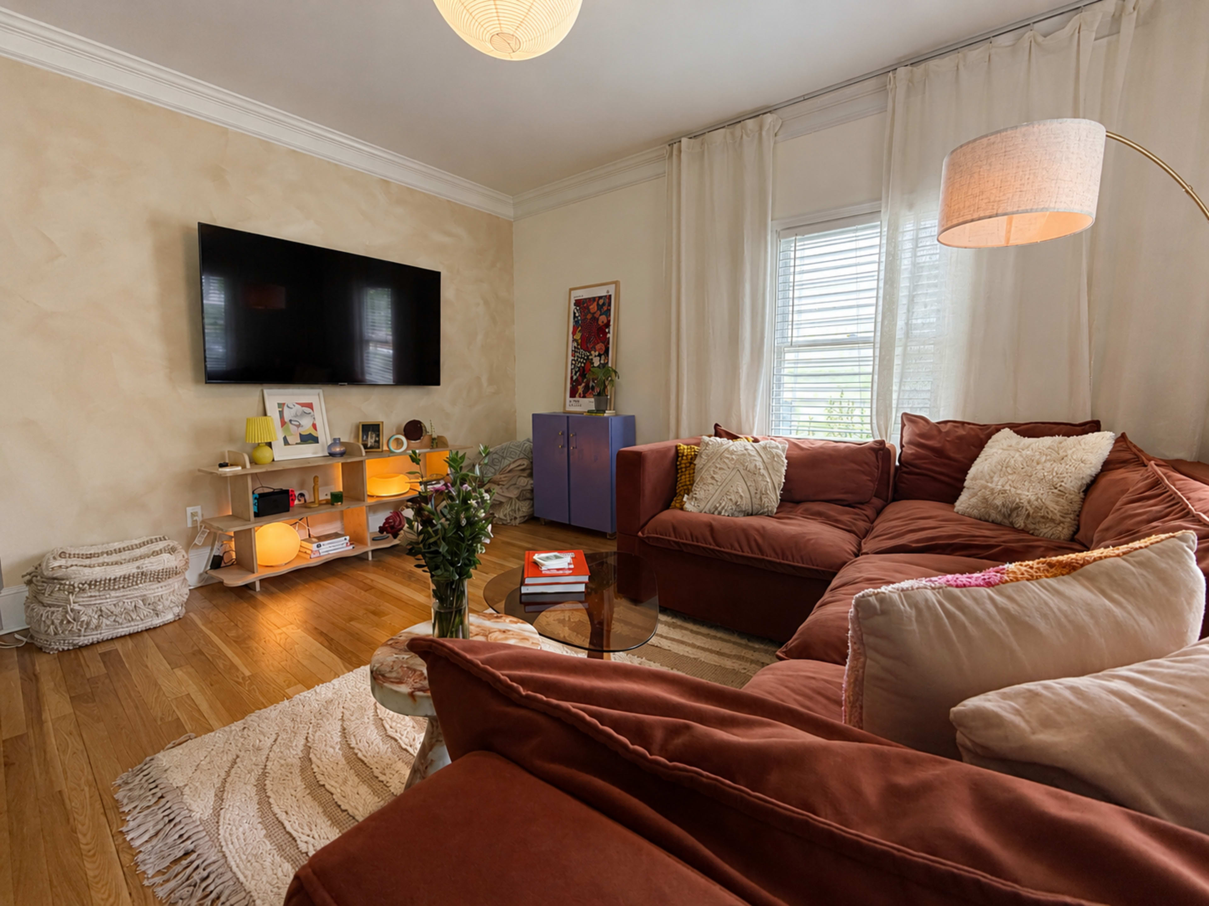 The image shows a cozy living room with a large sectional sofa, a flat-screen TV mounted on the wall, and a coffee table surrounded by decorative items and plants.