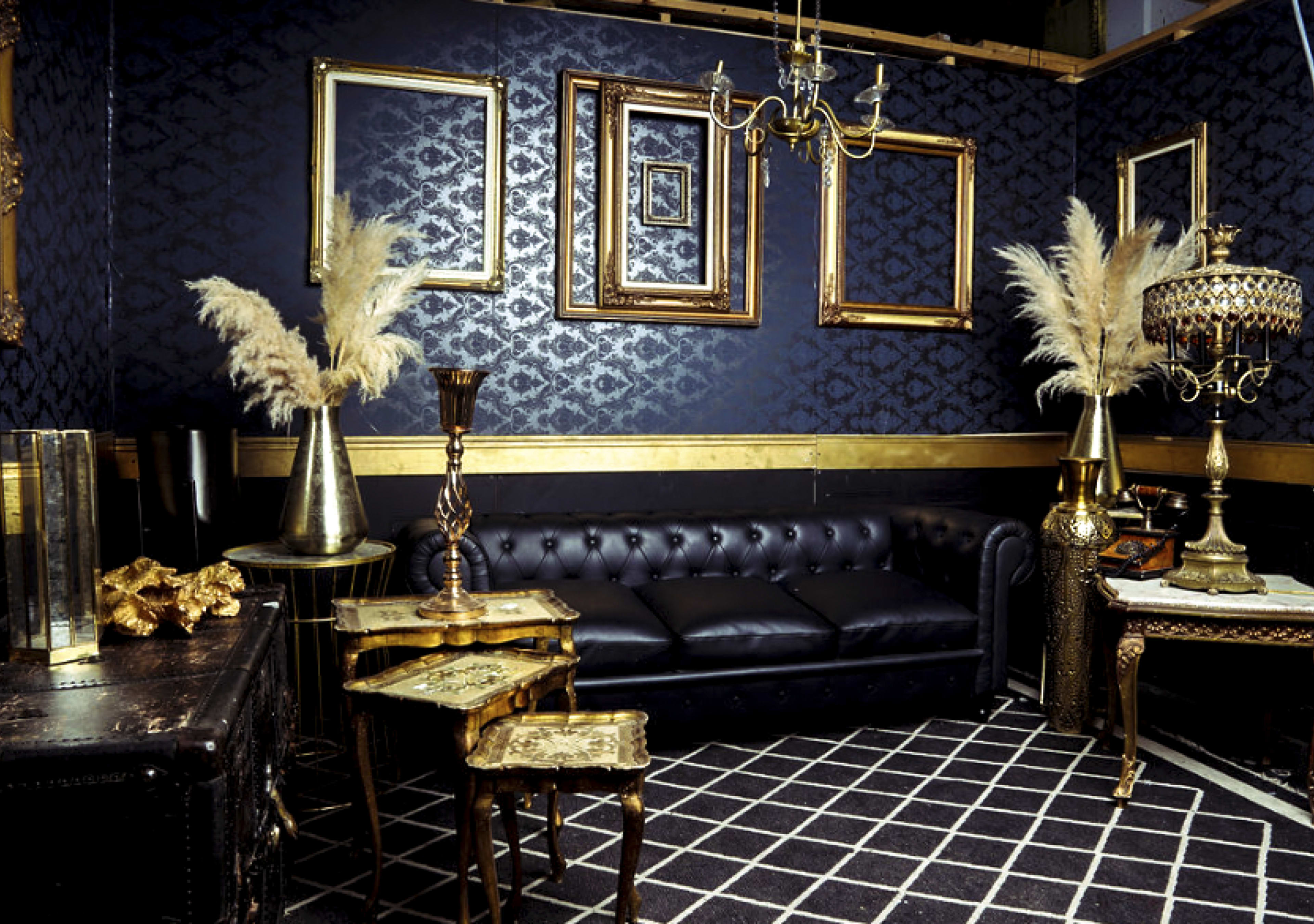 A dark, elegant room featuring a black leather couch, ornate furniture, and decorative gold-framed mirrors against a patterned wall.