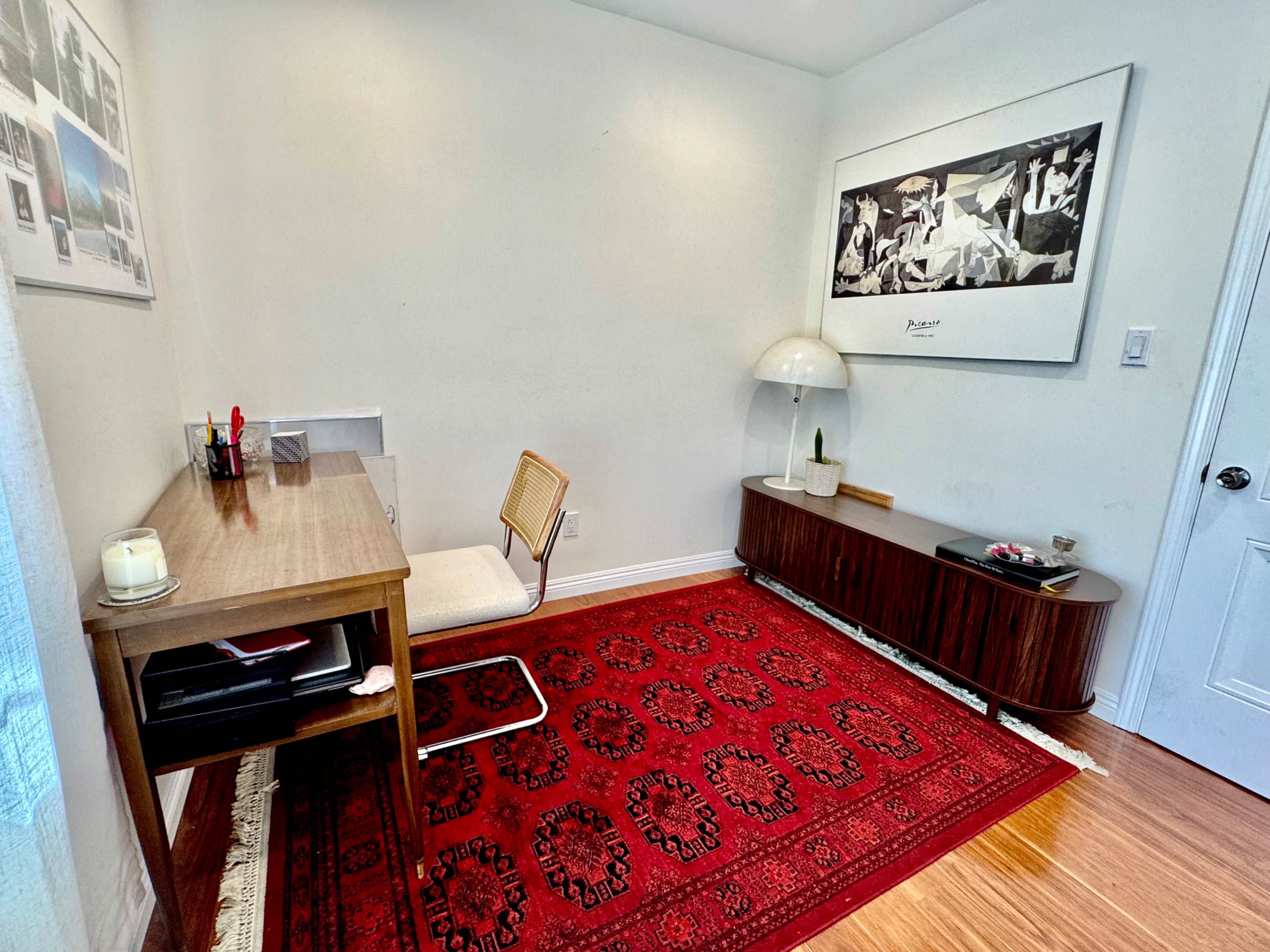 The image shows a minimalist office space featuring a wooden desk with a chair, a decorative rug, and a large framed artwork on the wall.