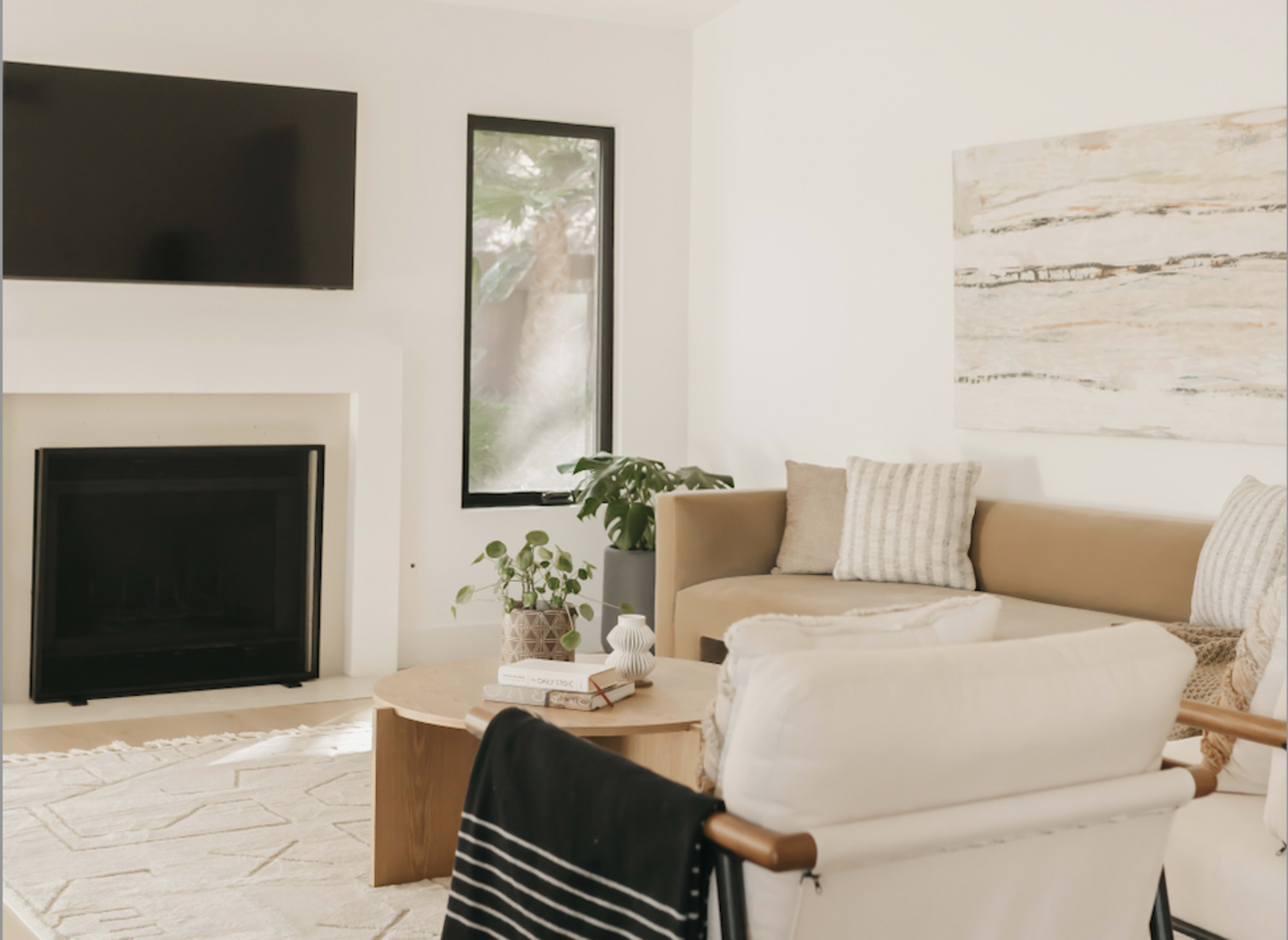 A modern living room featuring a beige sectional sofa, a round wooden coffee table, a large flat-screen TV mounted on the wall, and a plant near the window.