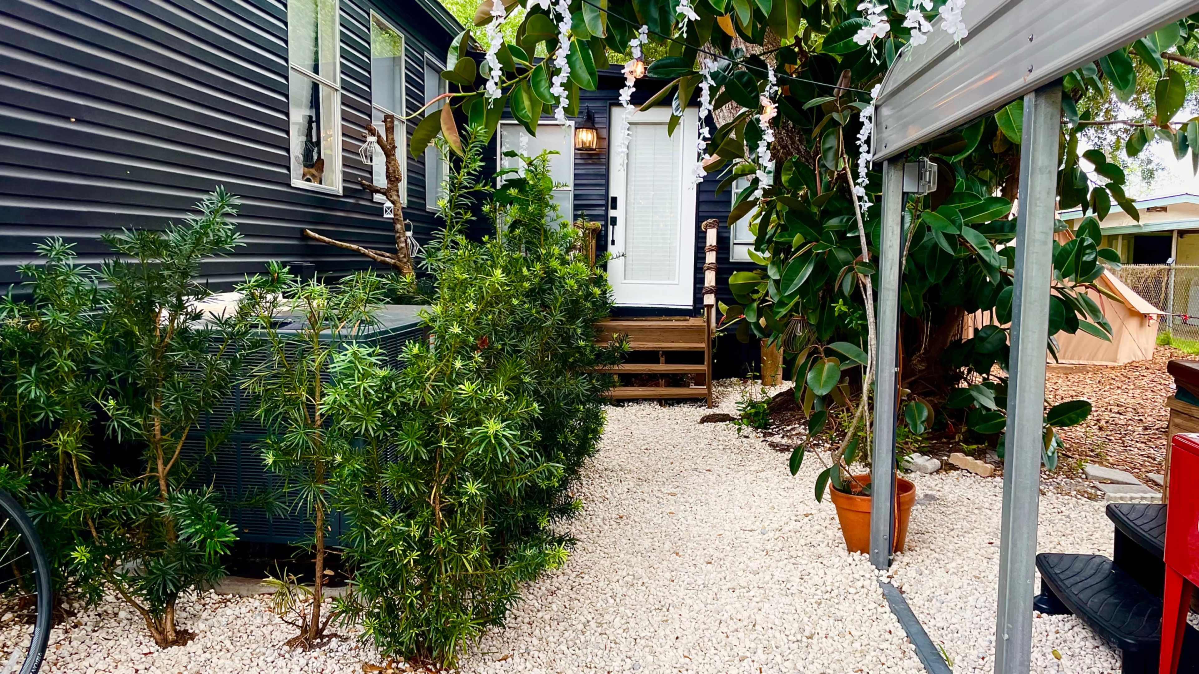 A narrow pathway lined with white gravel leads to a doorway surrounded by greenery and outdoor decorations.
