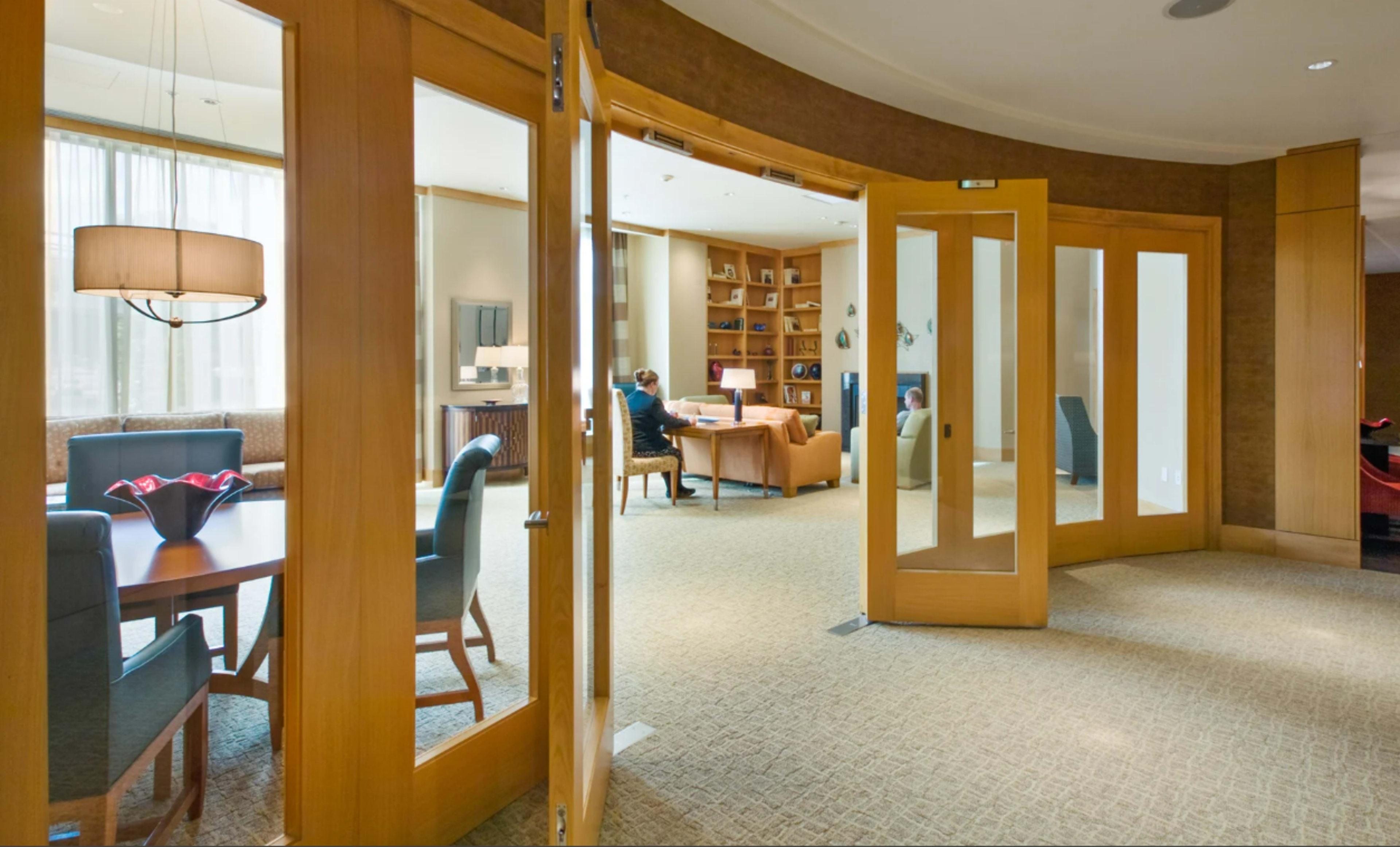 A spacious lounge area features wooden doors leading into a well-lit room with a seating area and a person working at a desk.