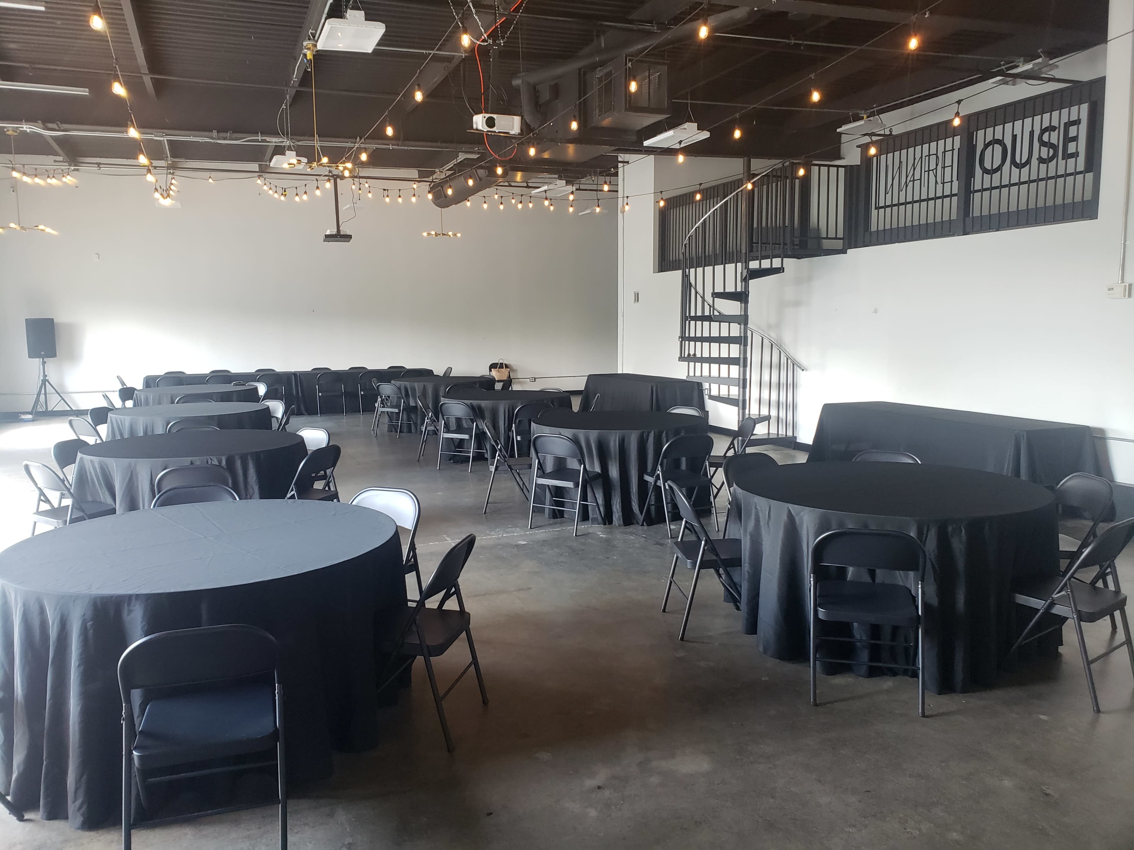A spacious event venue with several round tables covered in black tablecloths, arranged in a room with high ceilings and string lights.