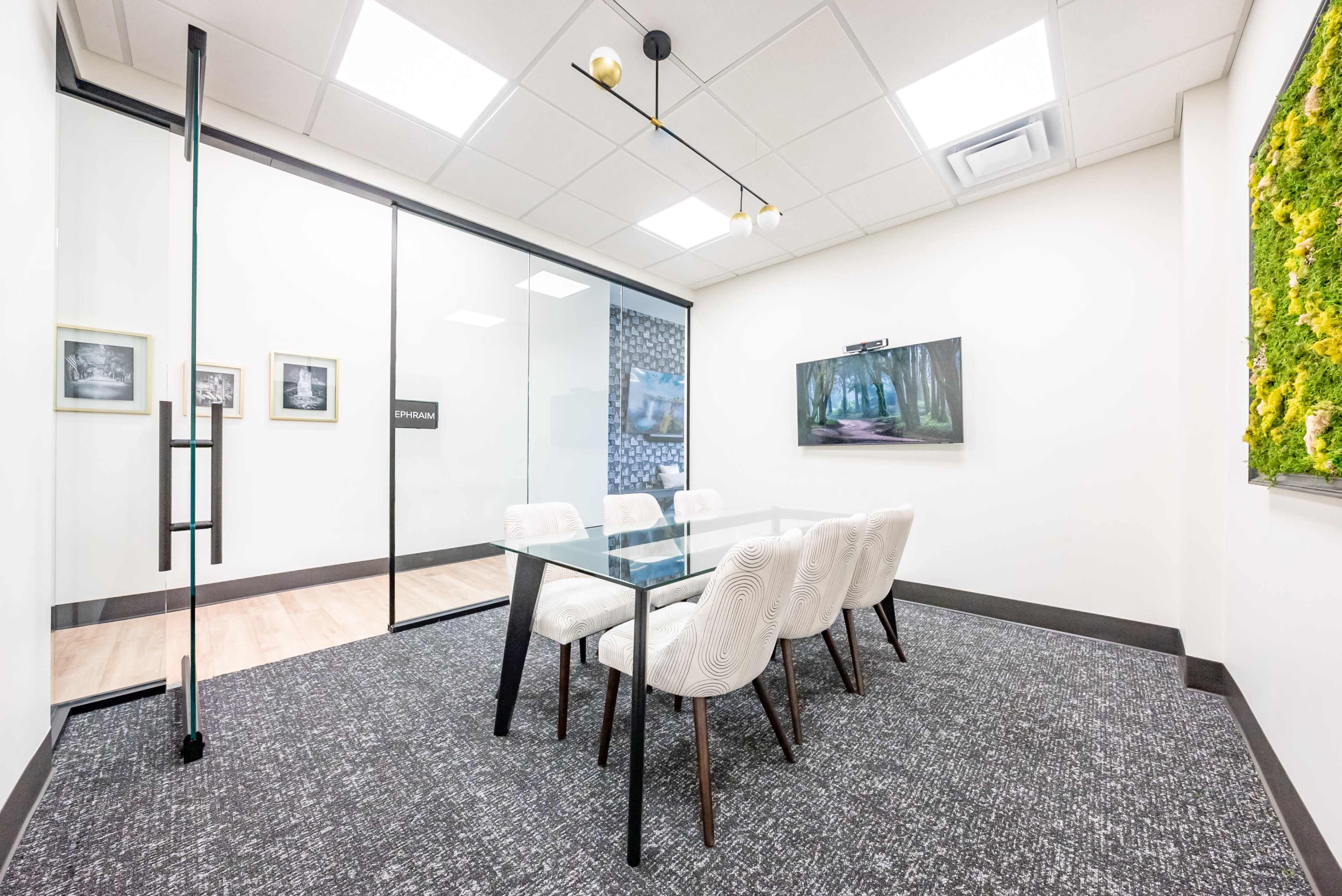 A modern conference room features a glass wall, a glass table with six upholstered chairs, and a large wall-mounted screen displaying a nature scene.