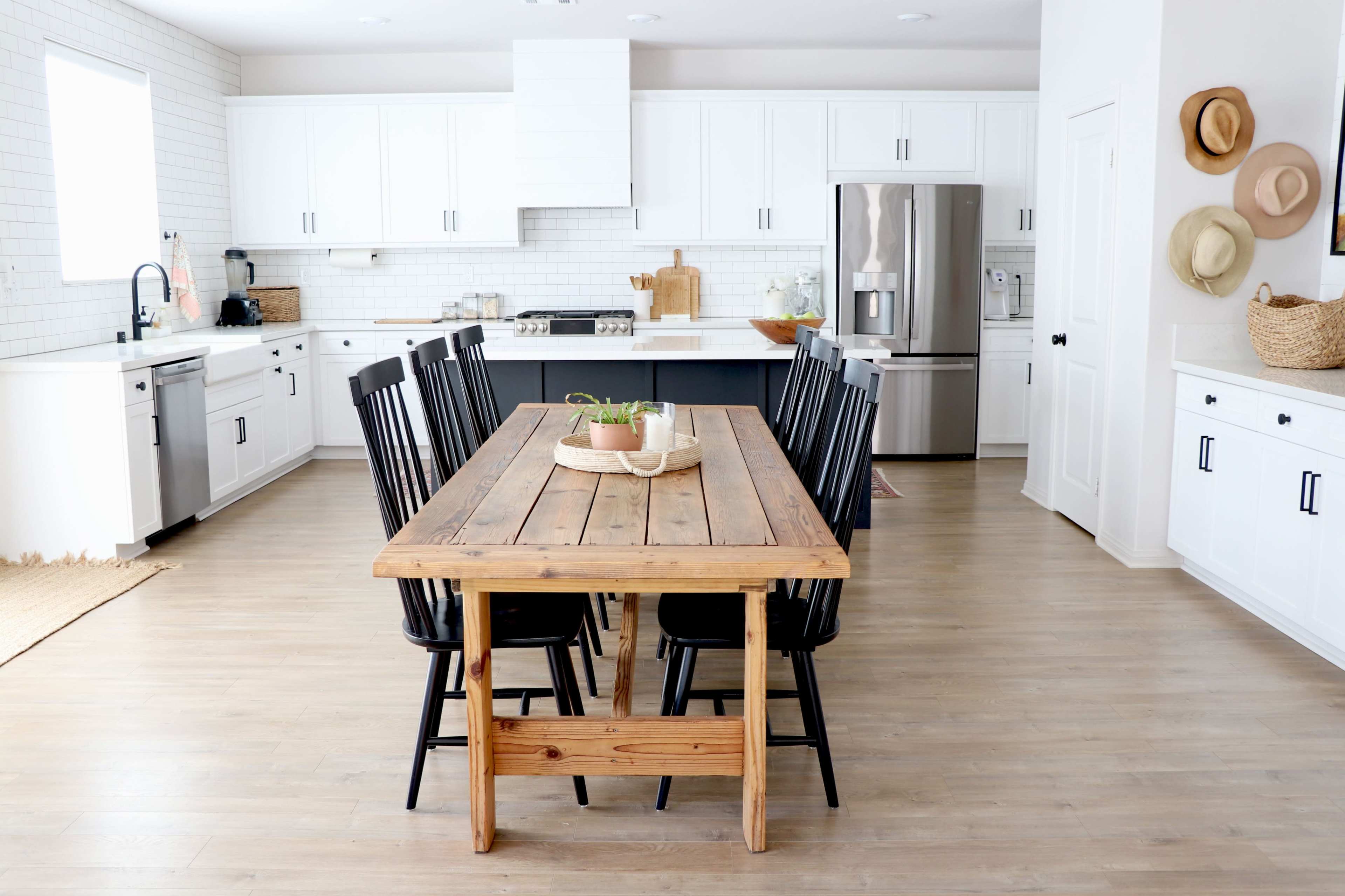 A large wooden dining table with black chairs is centered in a modern kitchen featuring white cabinets and stainless steel appliances.
