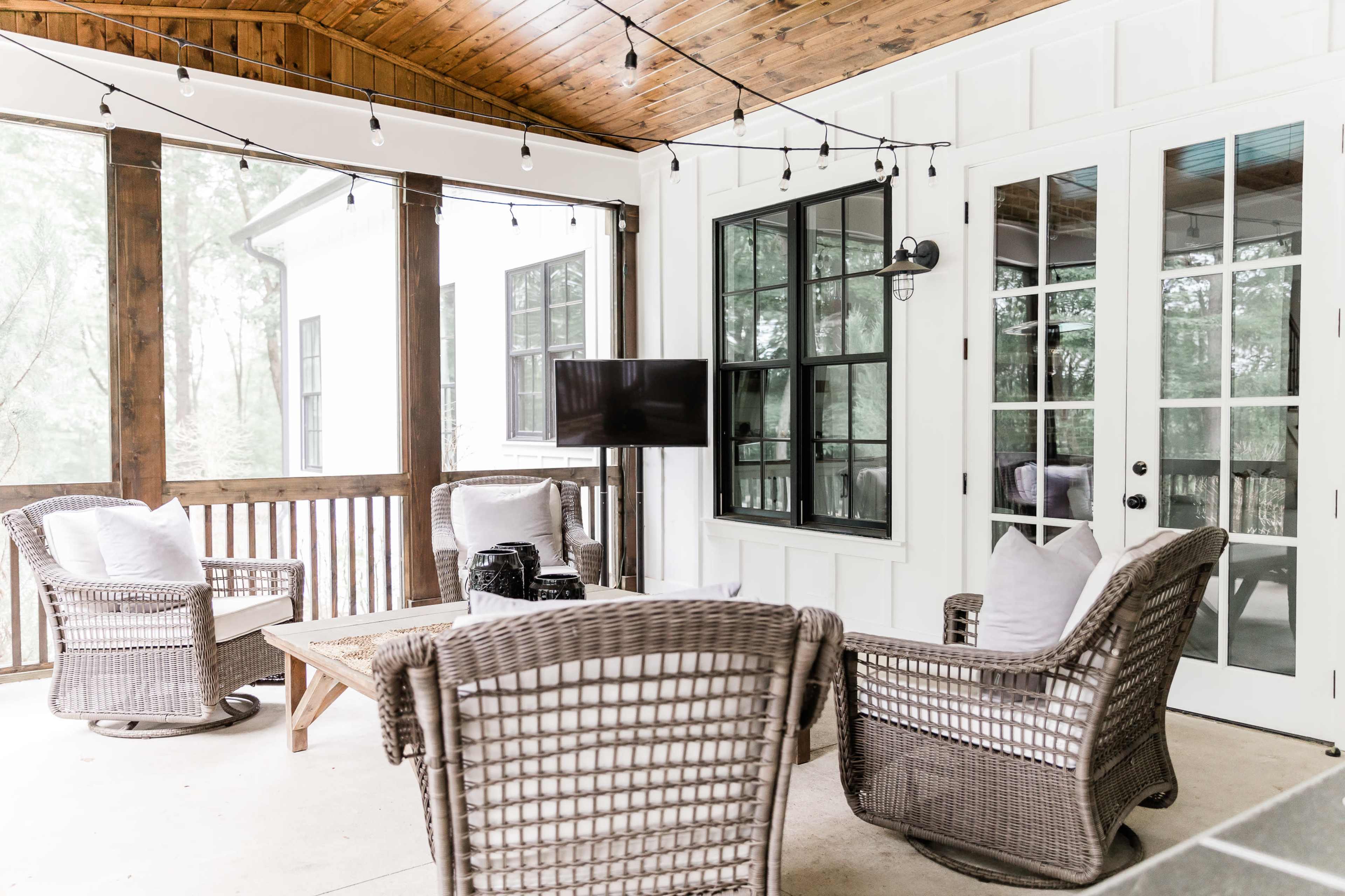 A screened-in porch with woven chairs, a coffee table, and a mounted television, all illuminated by string lights overhead.