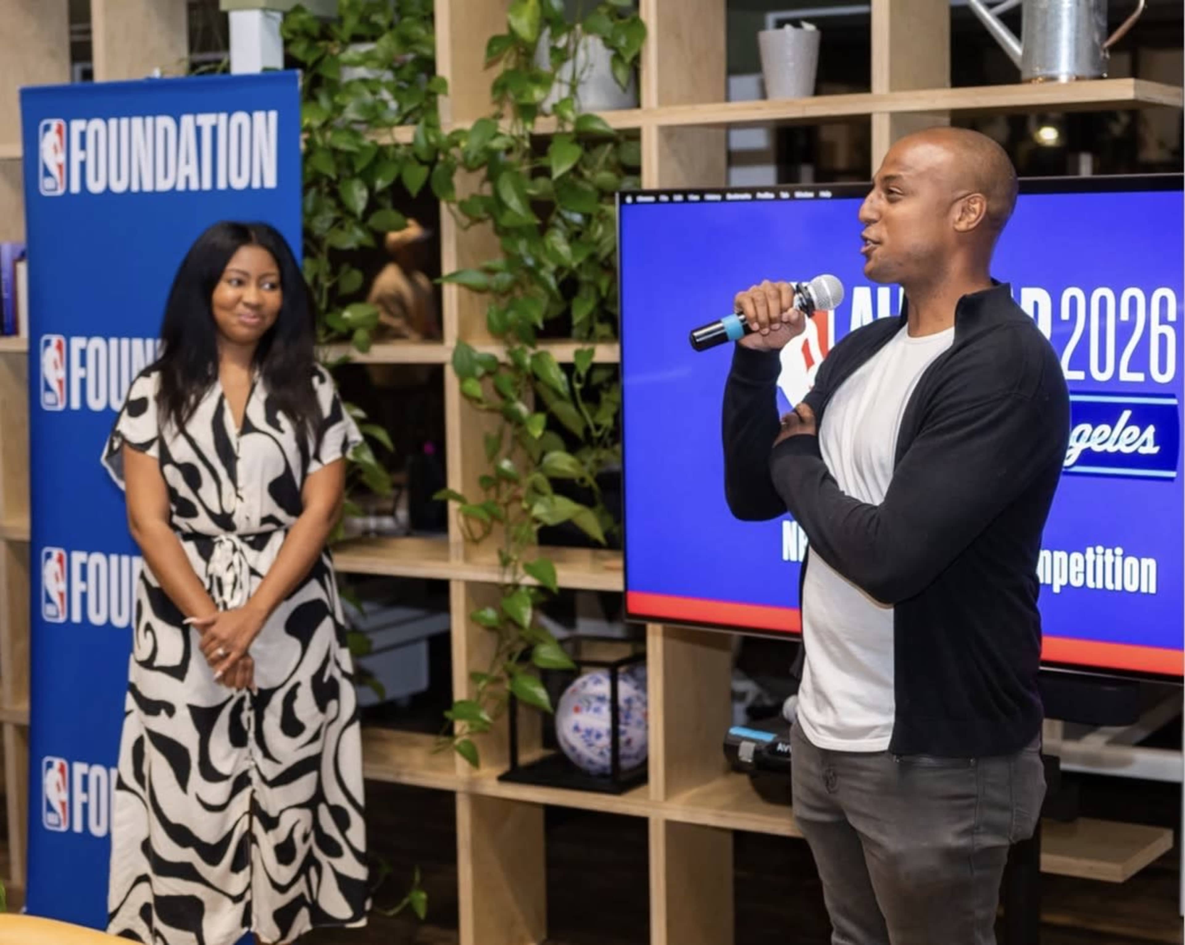 A man speaks into a microphone while a woman listens at an event featuring an NBA Foundation display in the background.