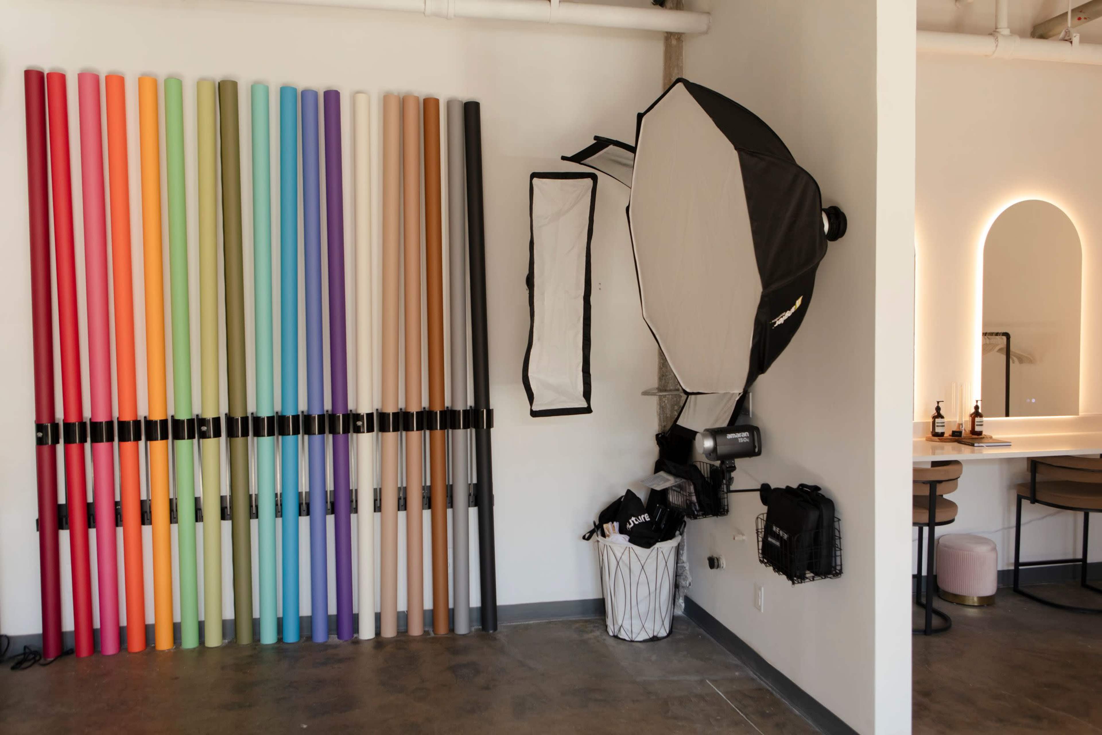 The image shows a well-lit studio with a wall of colorful backdrops on the left and photography equipment including a softbox on the right.