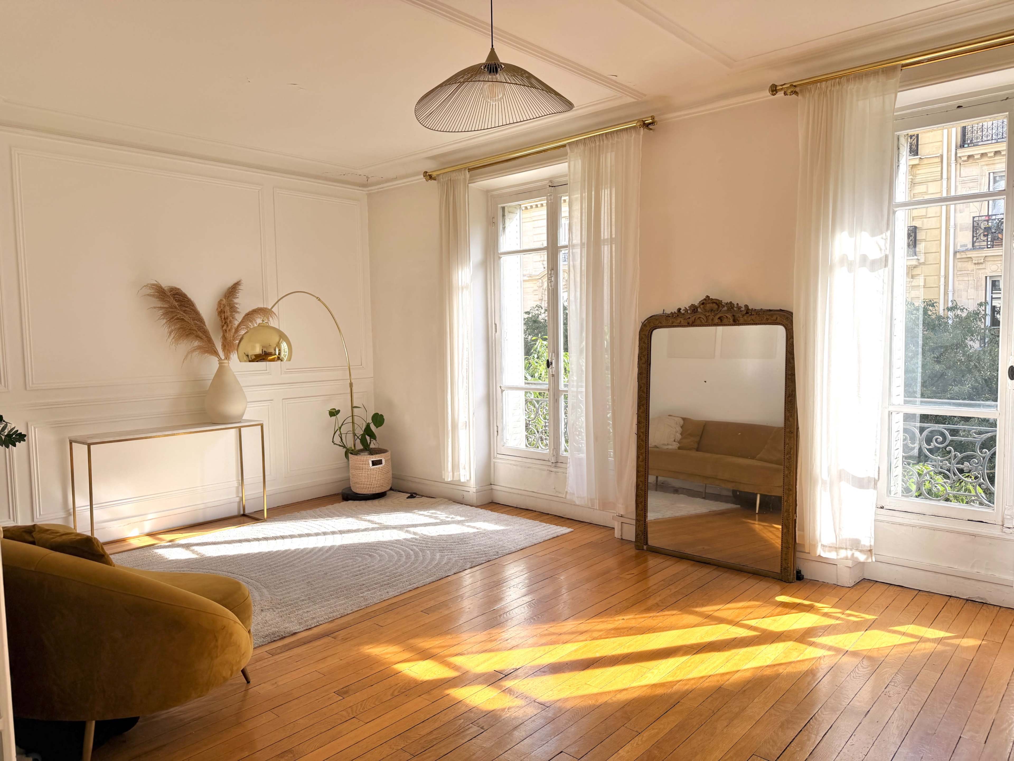 The image depicts a softly lit, minimalist living room with a large mirror, a comfortable sofa, and a plant, featuring wooden flooring and light curtains.