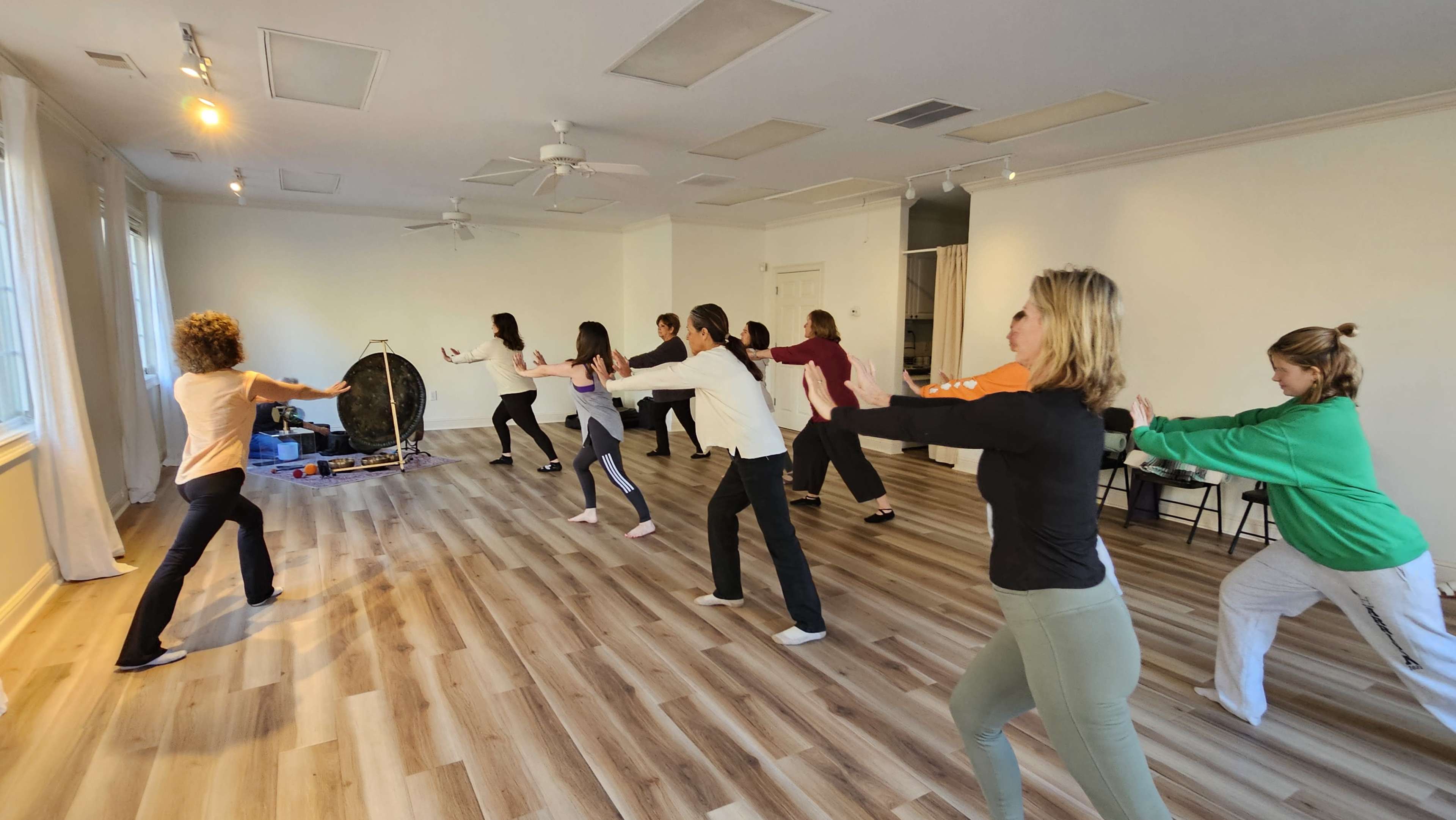 A group of people are practicing a coordinated movement exercise in a spacious, well-lit room with wooden flooring.