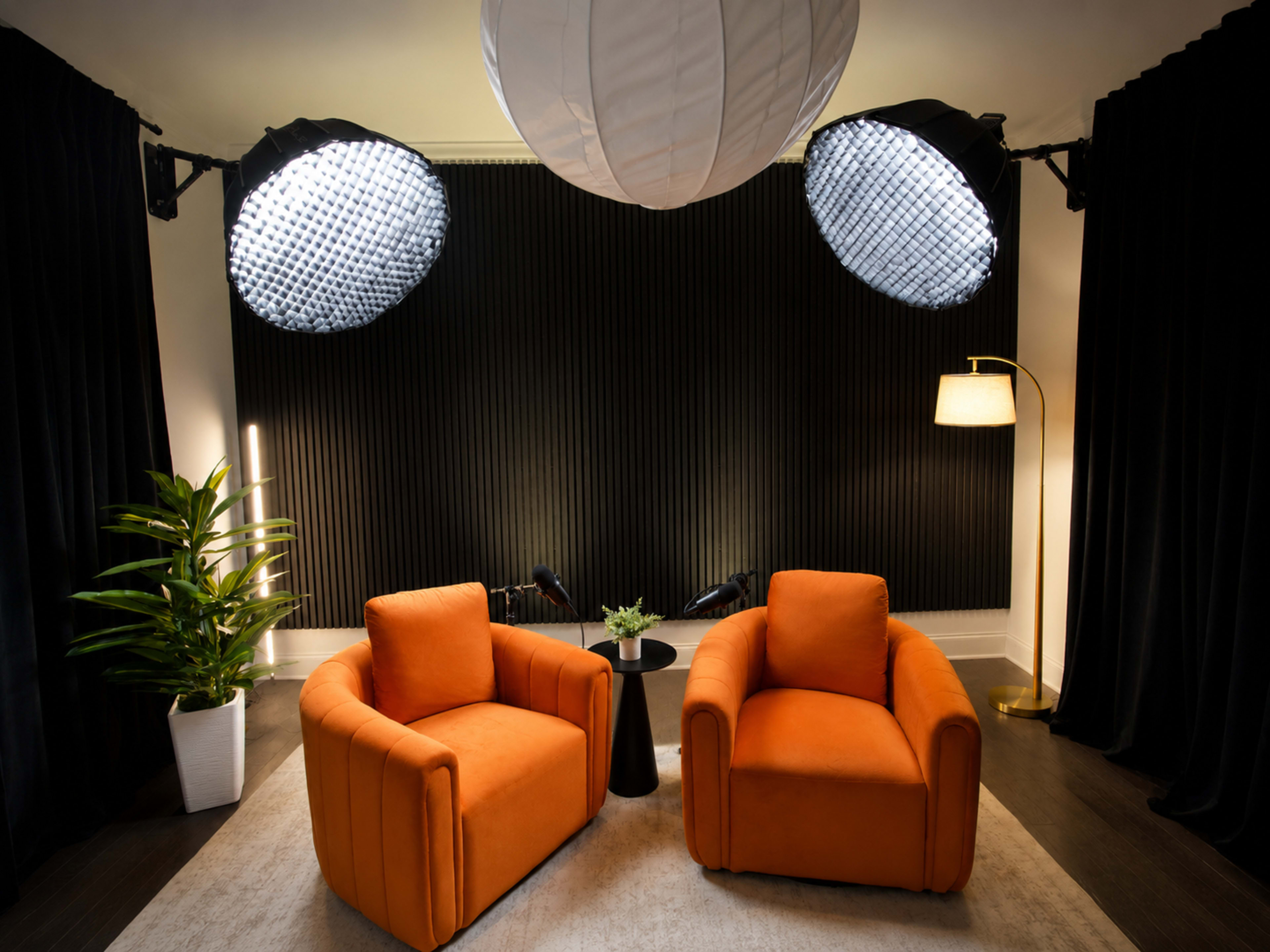 The image depicts a stylish indoor scene with two orange armchairs facing a small black table, illuminated by two large lights above and complemented by a decorative plant and lamp nearby.