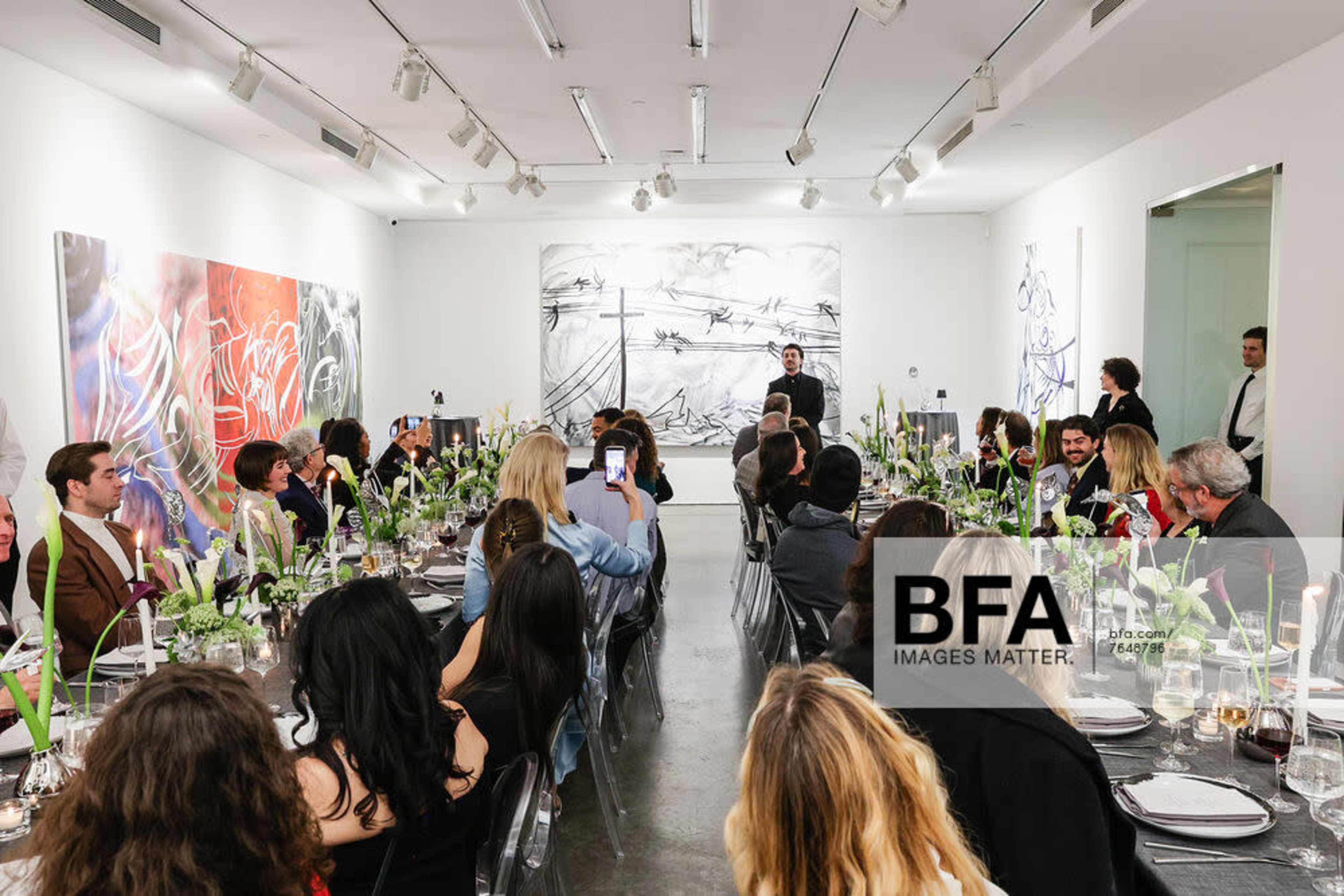 A large group of people sits at a long dining table in a gallery, with colorful artwork displayed on the walls and a speaker addressing the attendees.