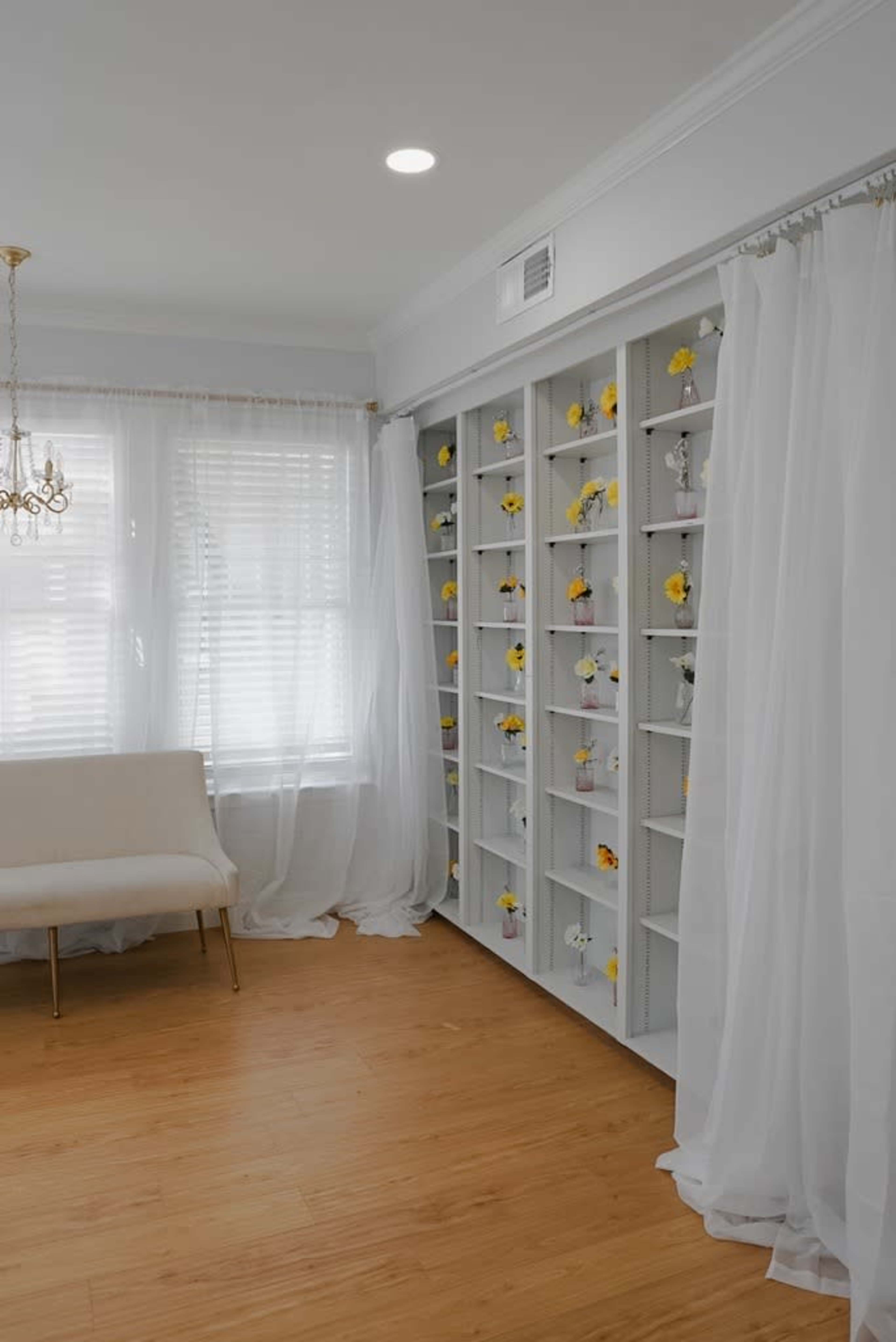 A bright room features a pale sofa beside a wall of shelves adorned with small yellow flowers and sheer white curtains.