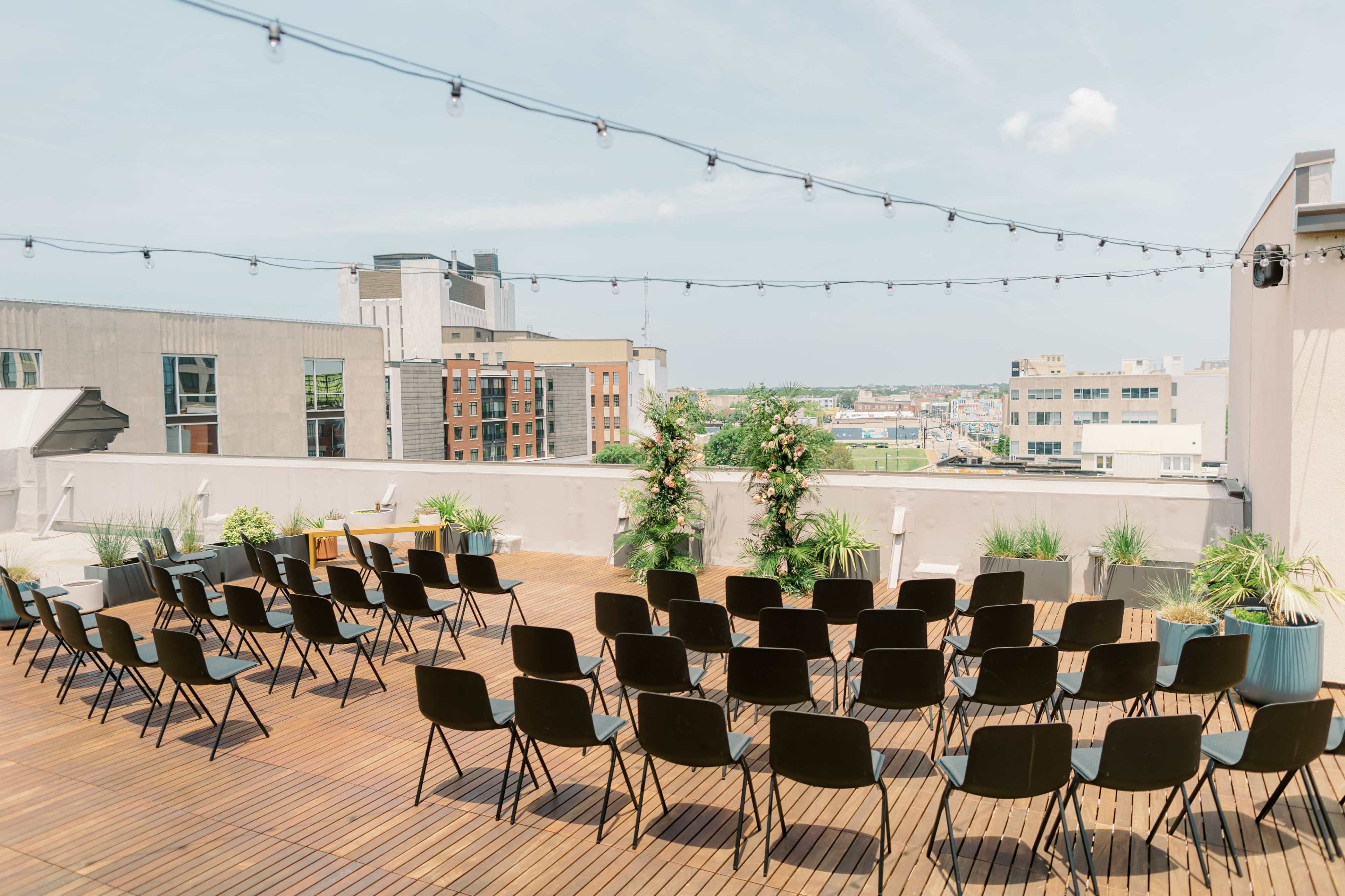 The image shows a rooftop venue set up with rows of black chairs arranged on a wooden deck, overlooking a city skyline under a clear sky.