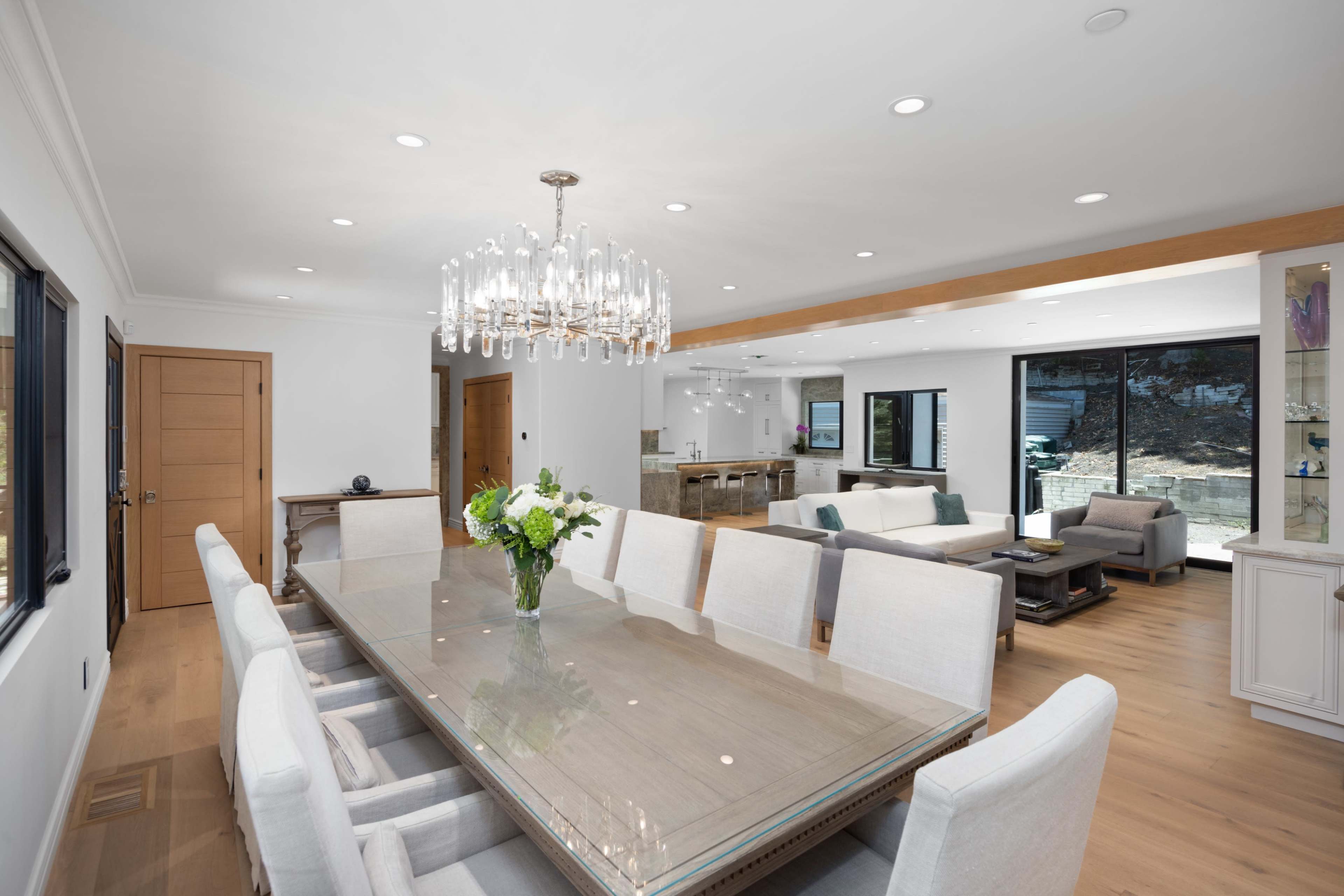 A modern dining area features a long glass table surrounded by upholstered chairs, with a chandelier overhead and large windows showcasing an outdoor view.
