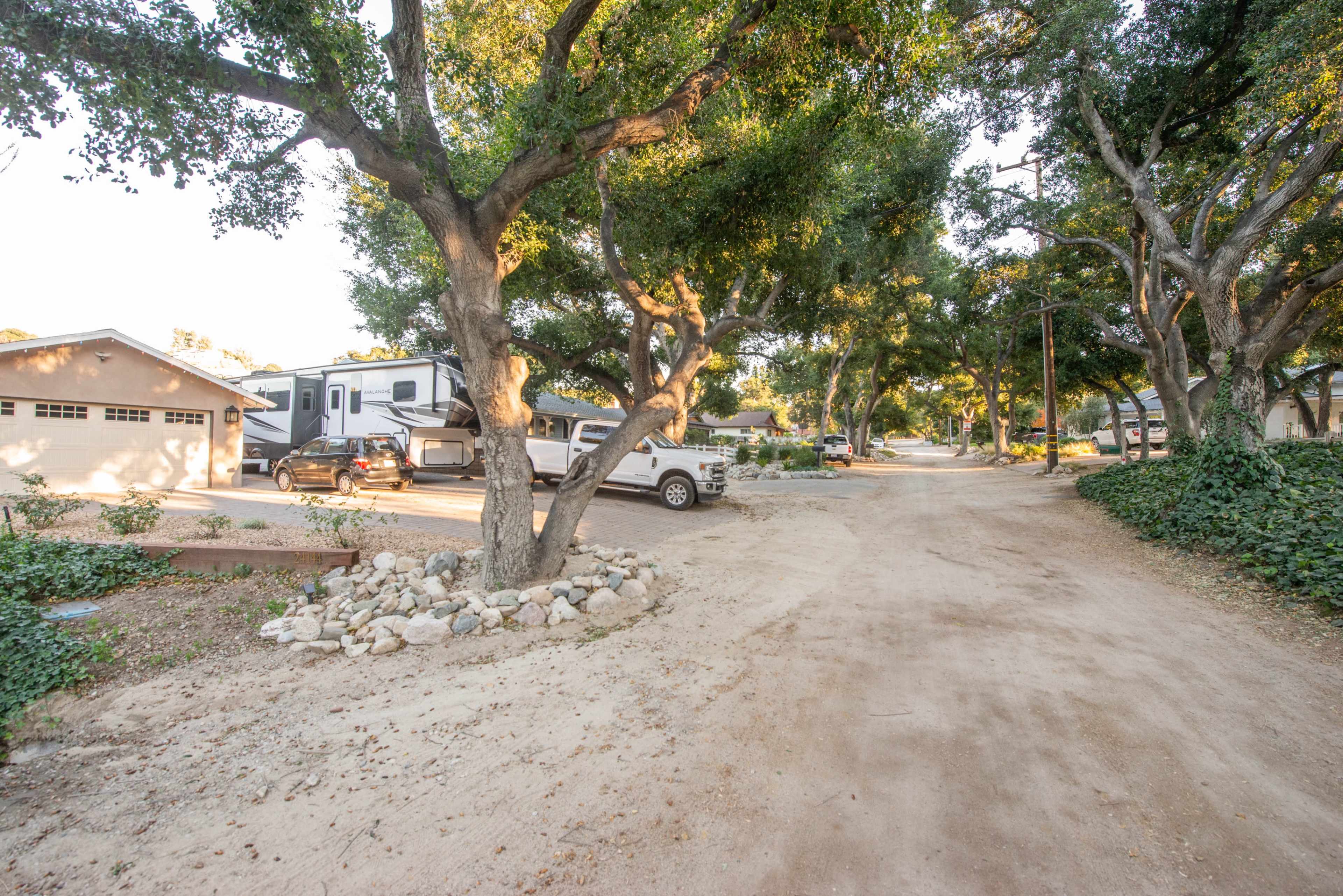 A dirt road lined with trees leads to parked vehicles near residential buildings.
