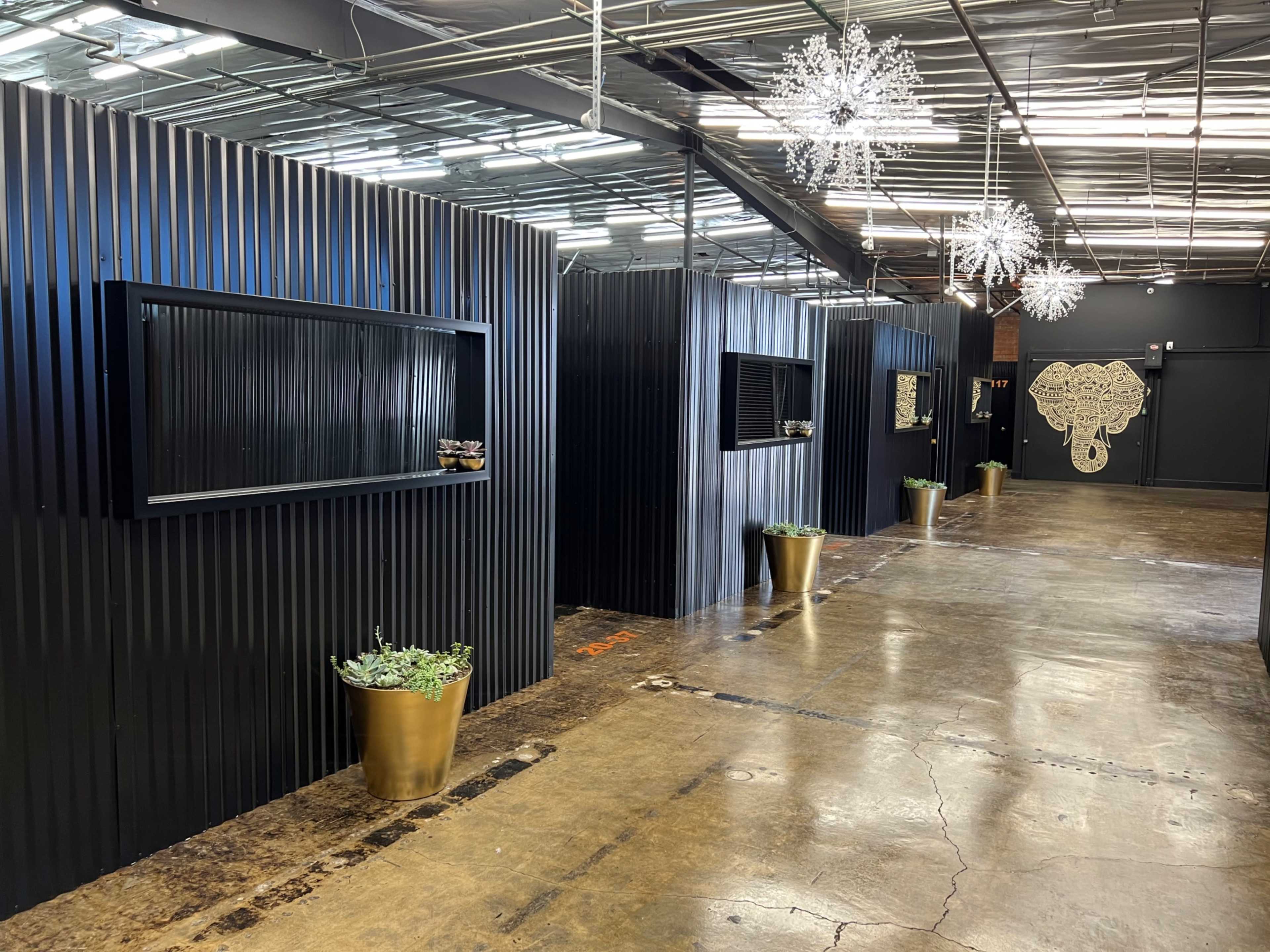 The image shows a modern indoor space with black corrugated walls, decorative mirrors, potted plants, and a whimsical elephant mural at the far end.