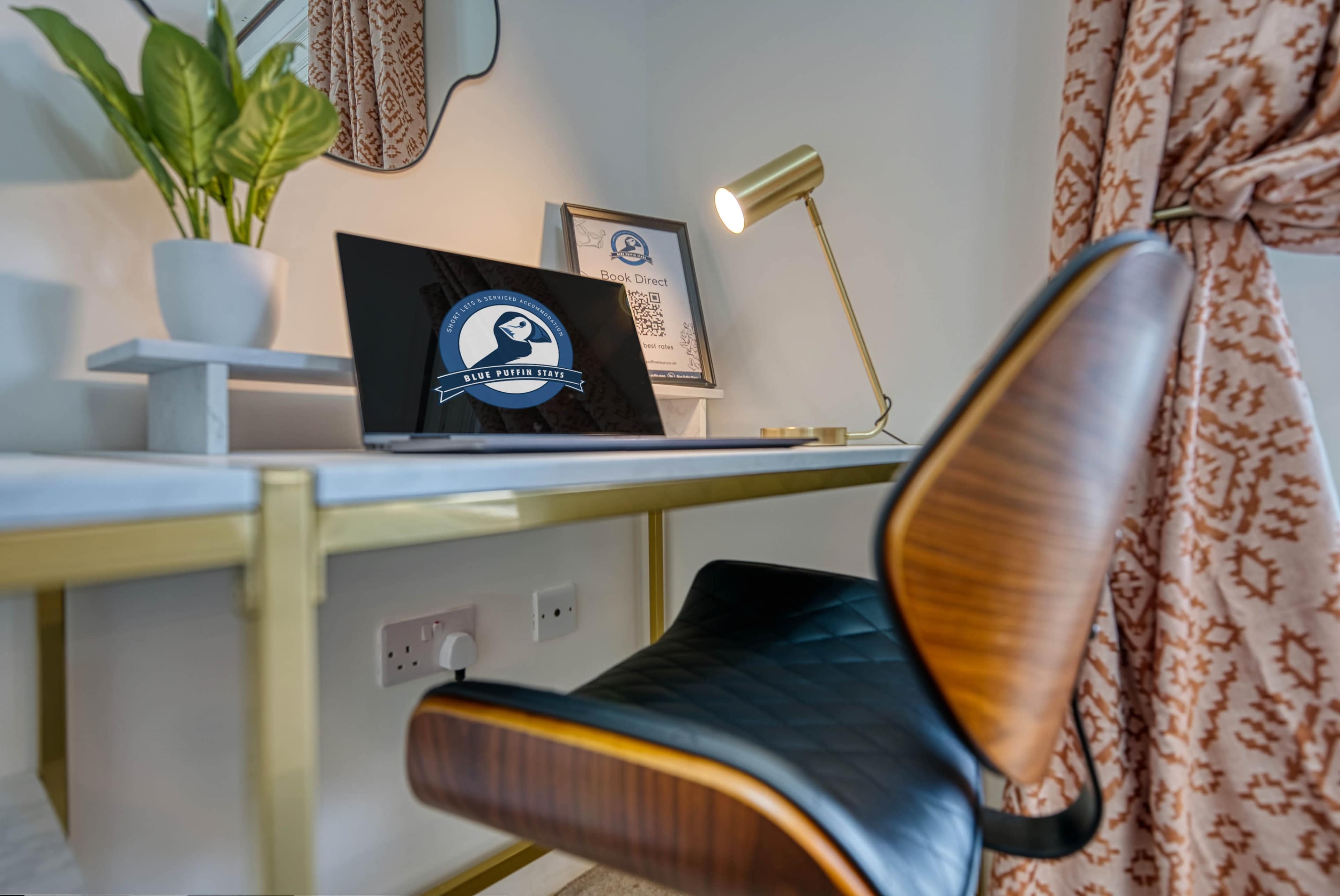 A modern desk with a laptop, a stylish lamp, a potted plant, and decorative items is shown in a well-lit room.