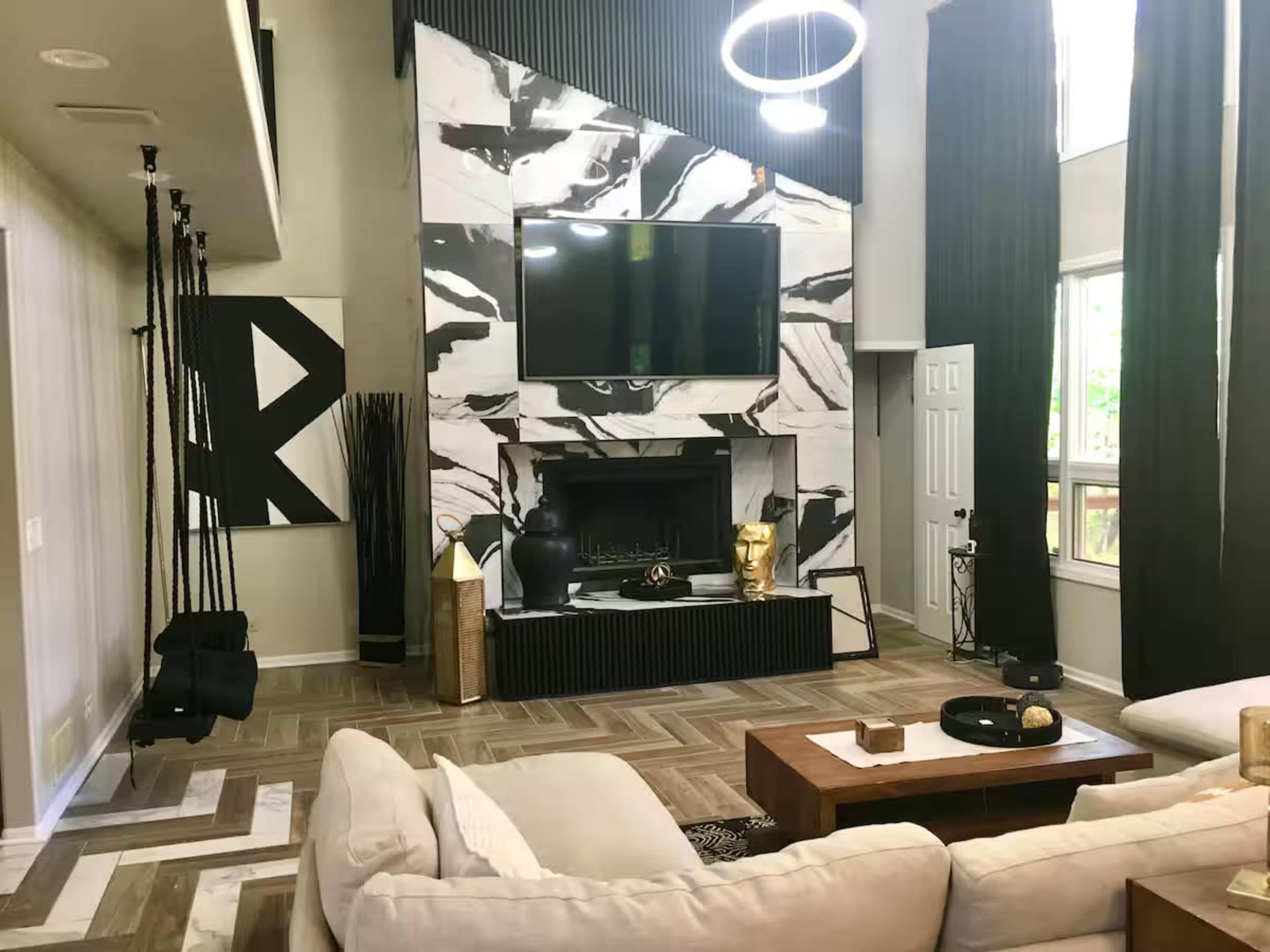 The image shows a modern living room featuring a large abstract-patterned fireplace, a mounted television, a sectional sofa, and various decorative elements, including plants and sculptures.