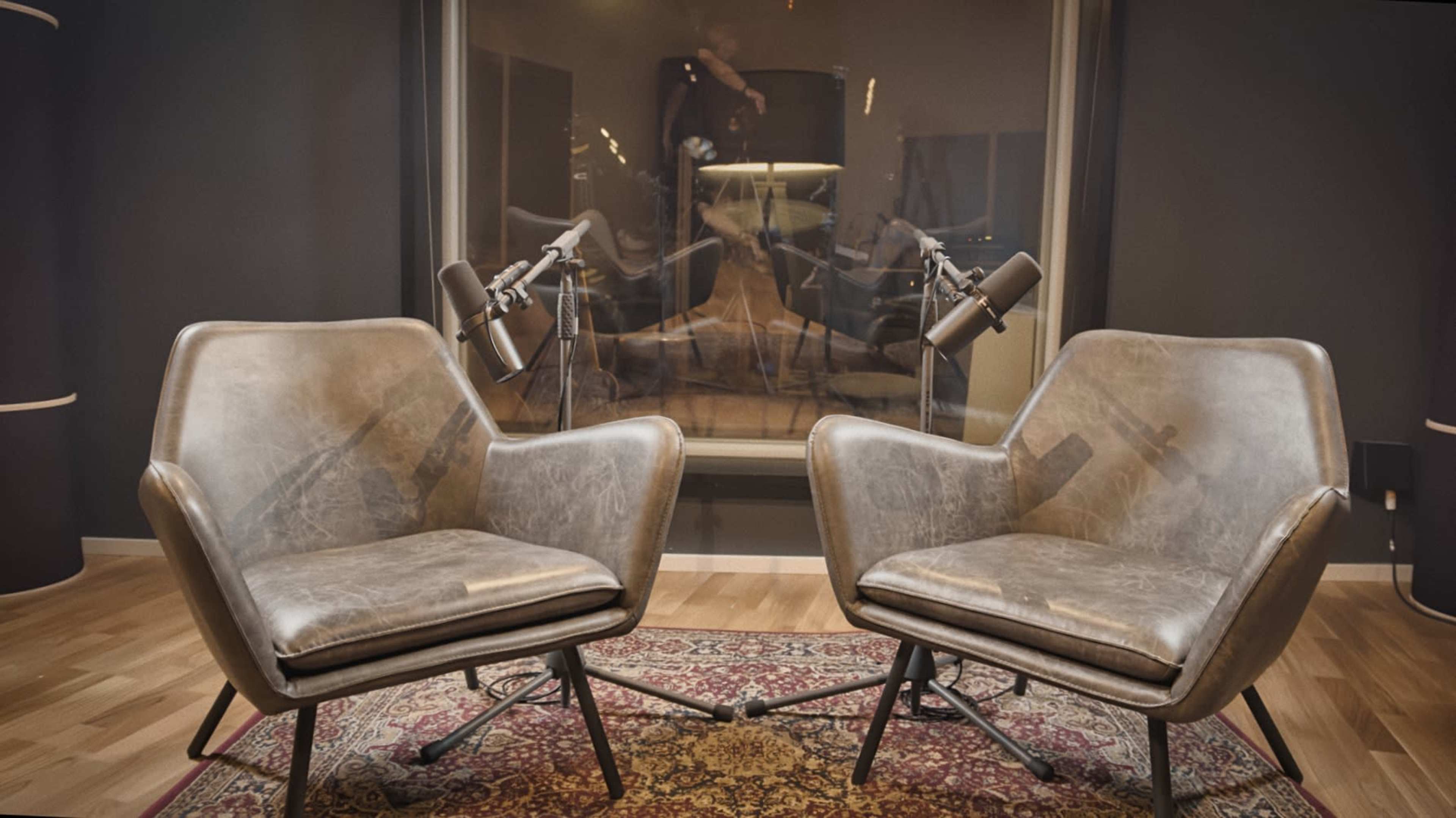Two modern chairs with a leather finish are positioned on a patterned rug in front of a soundproof glass window, which reflects two microphones set on stands.