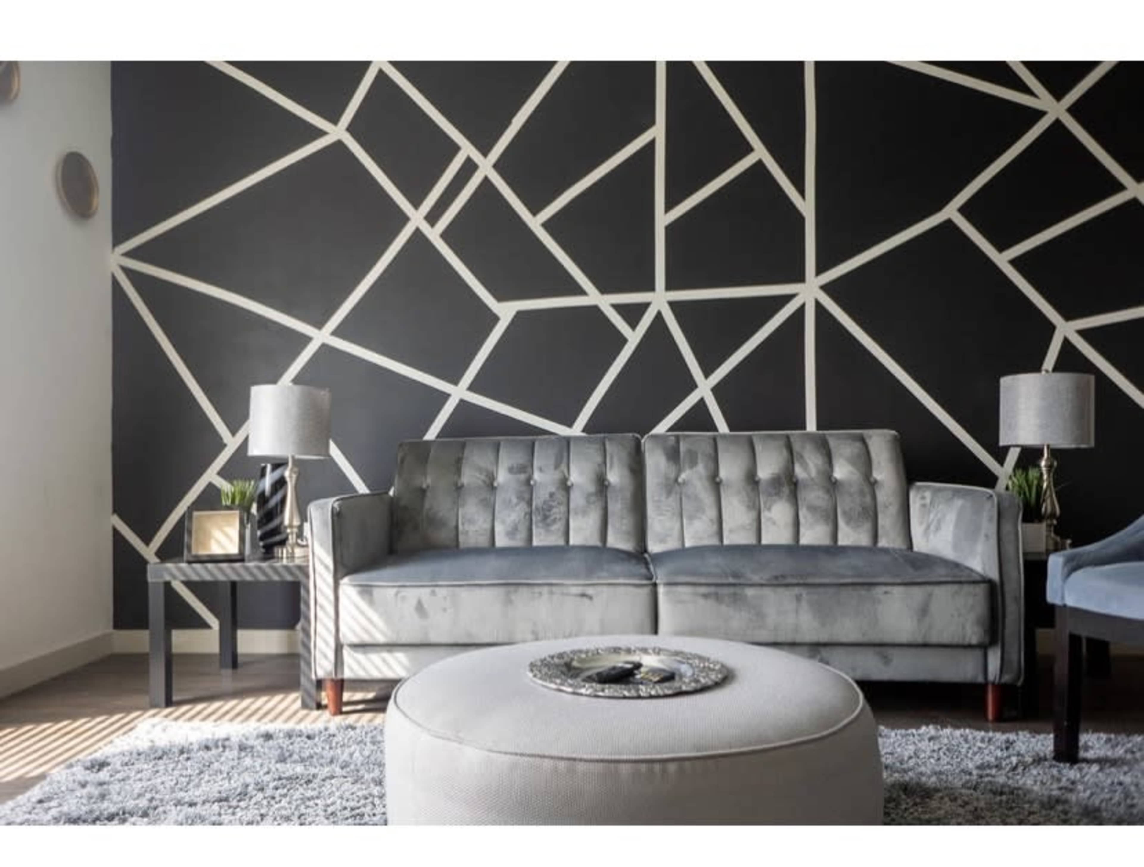 A modern living room with a gray velvet sofa, a round ottoman, and a geometric black and white accent wall.