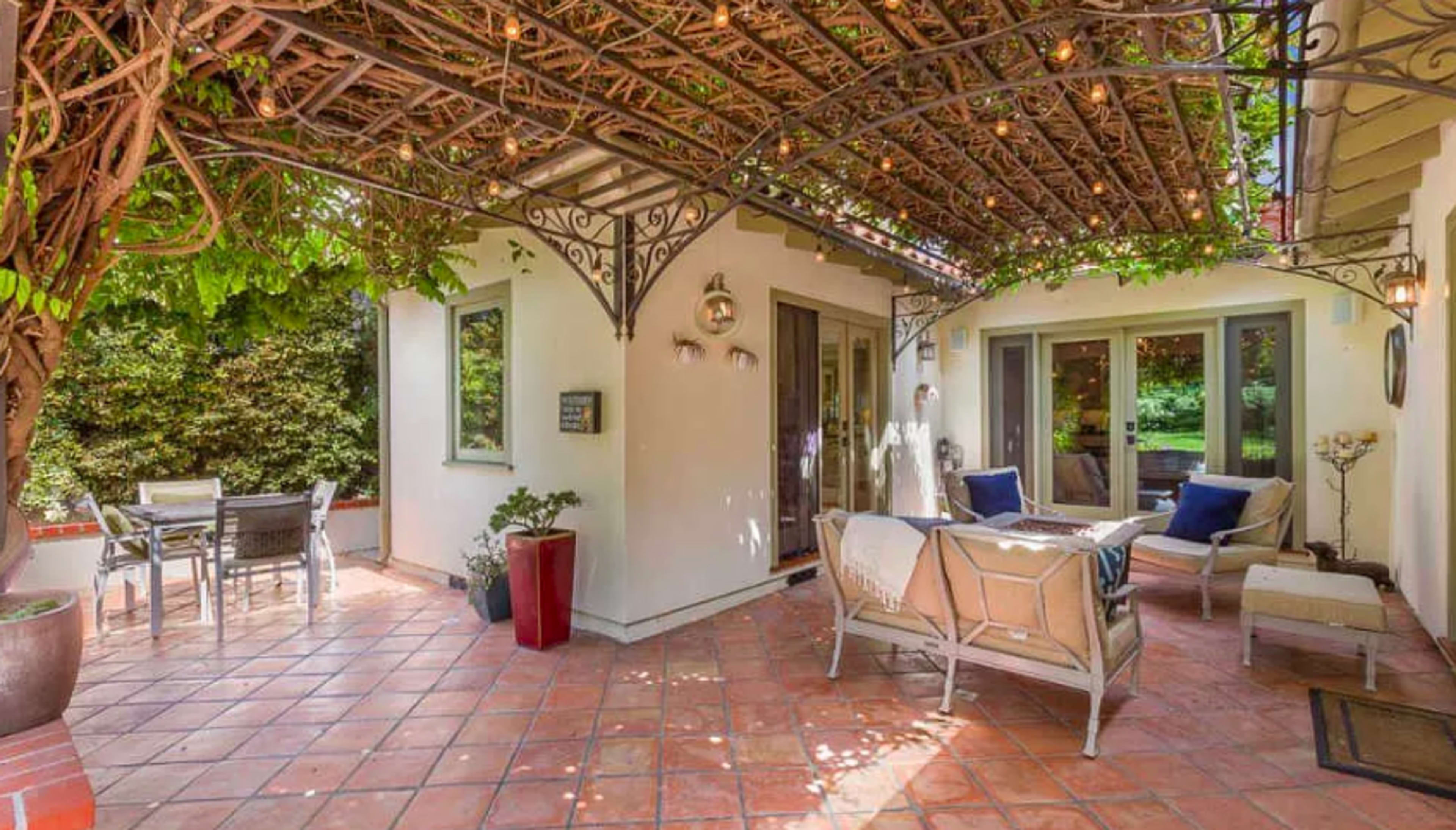 The image features a patio area with a seating arrangement and a dining table, all shaded by a trellis covered in greenery.