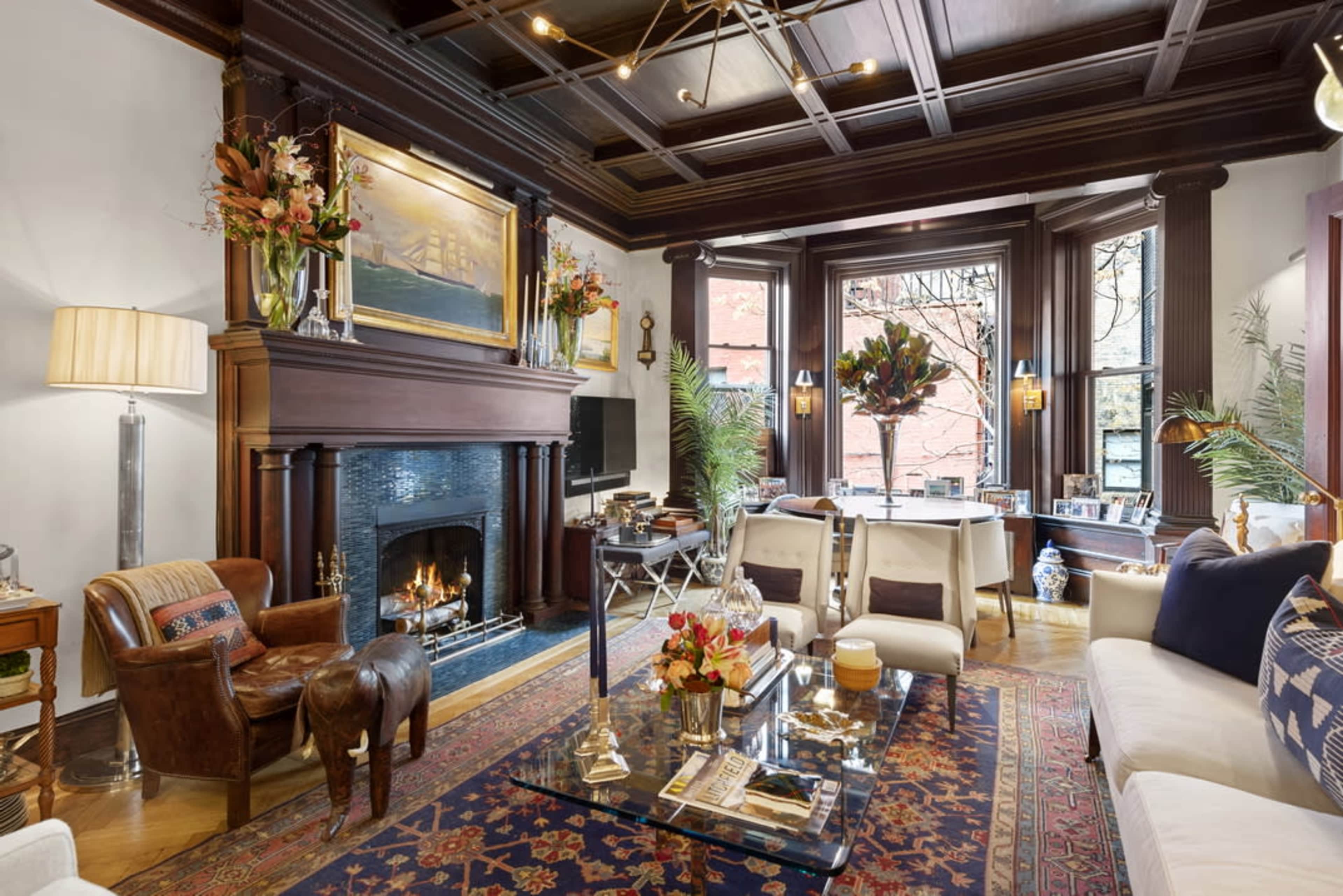 The image shows a richly decorated living room featuring a fireplace, a large window with views of greenery, and elegant furniture arranged around a glass coffee table.